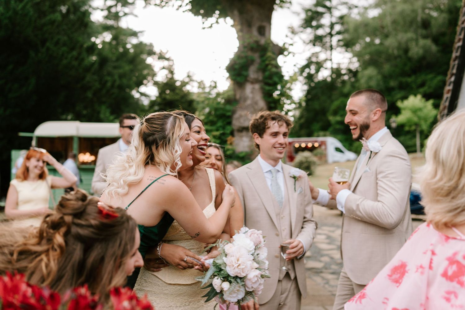 Bride hugging a guest while groom smiles at an outdoor wedding reception surrounded by trees.