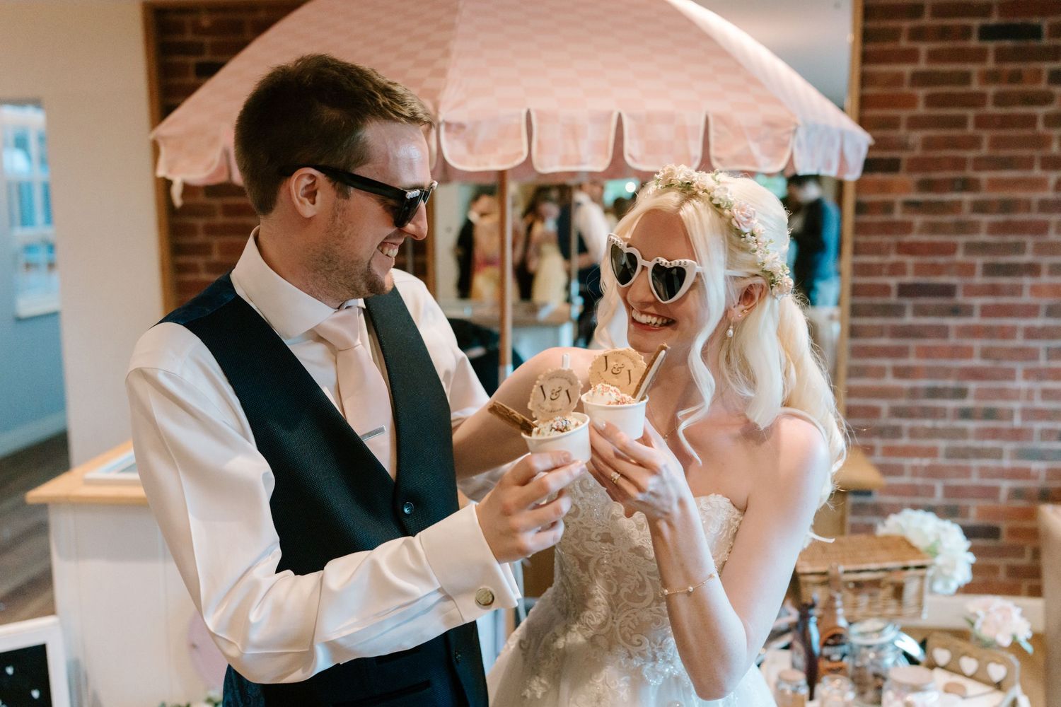 Bride and groom sharing ice cream cups at wedding reception, wearing sunglasses and smiling under pink umbrella.