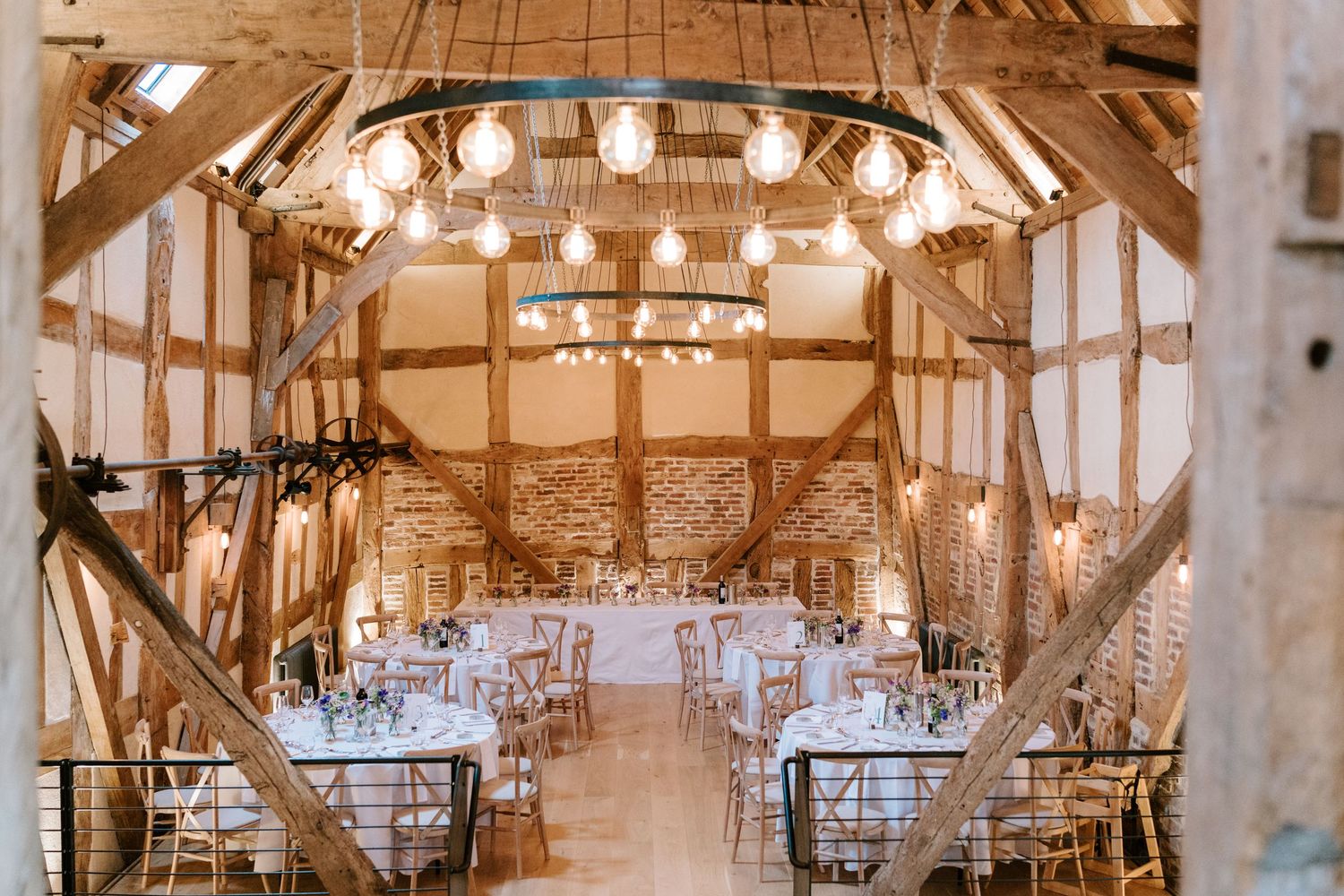 Rustic barn wedding reception venue with exposed timber beams, hanging Edison bulbs, and elegantly set round tables.