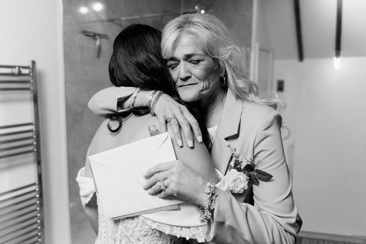 Two women share an emotional hug at a wedding, one holding a card, captured in black and white photography.