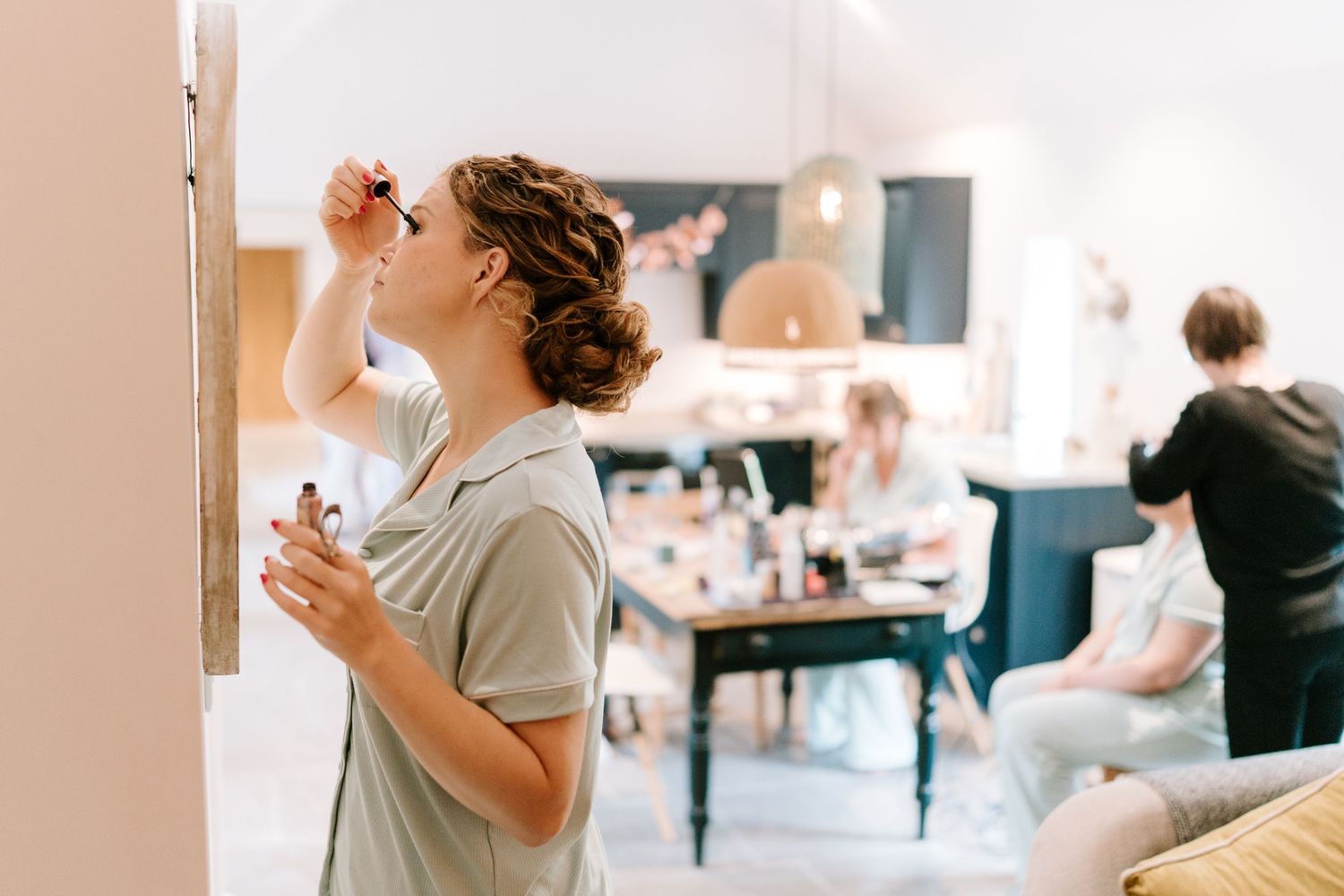 Woman applying mascara in mirror while others get ready in bright salon background.