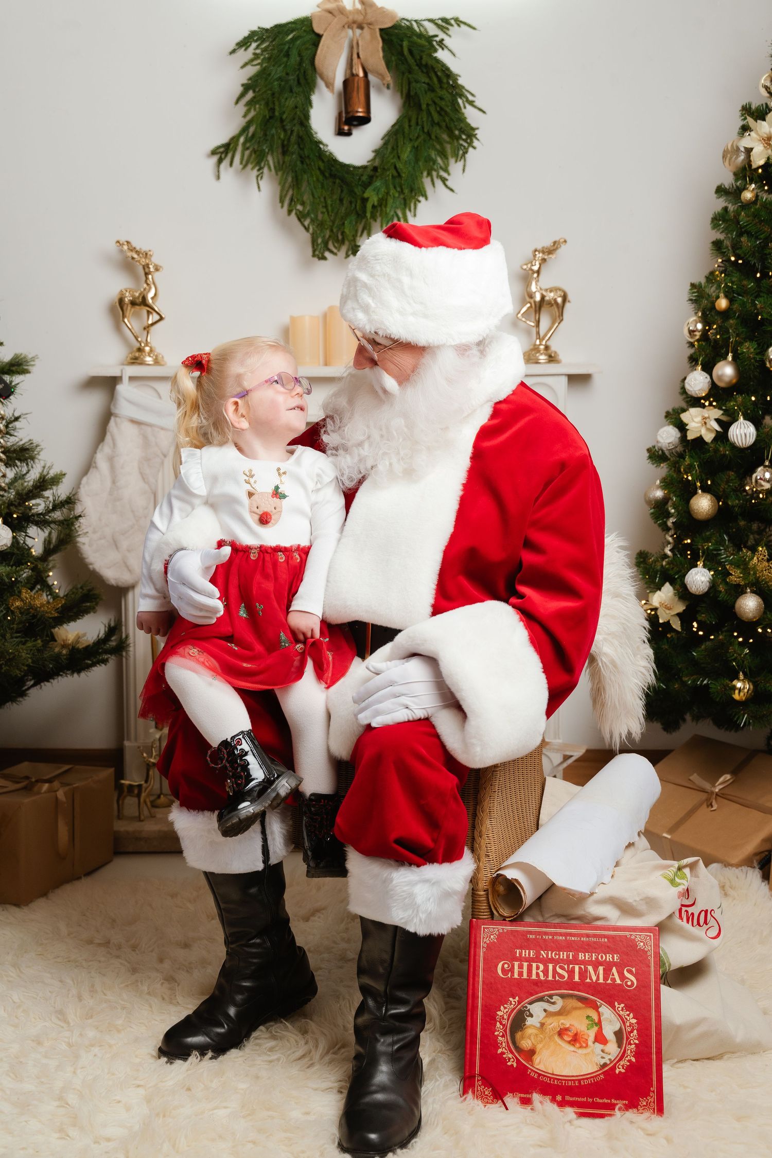 A little girl in red dress sits on Santa Claus lap beside a Christmas tree and The Night Before Christmas book.