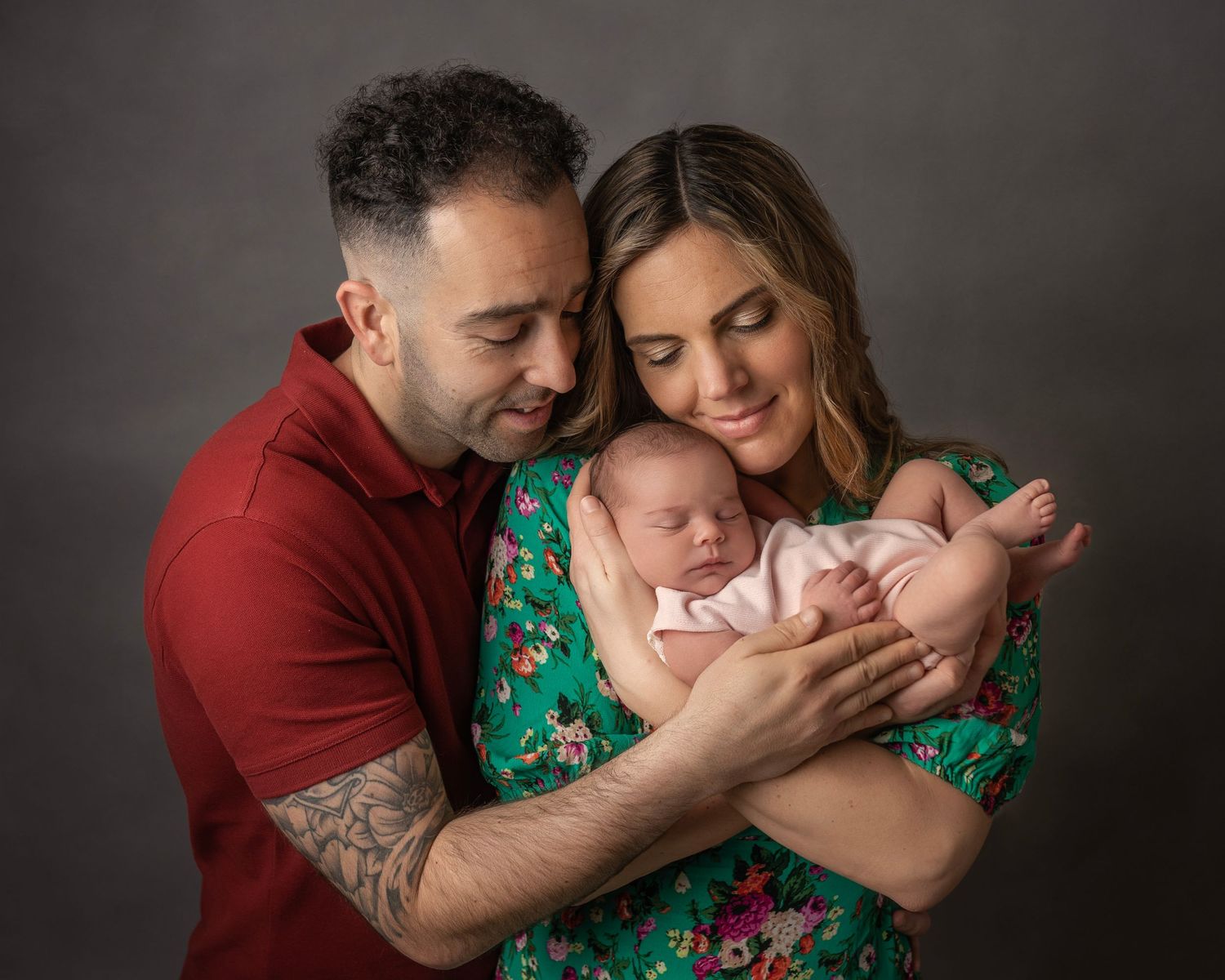 A couple tenderly holds their sleeping newborn baby, the mother in a floral dress and father in red, against a gray backdrop.