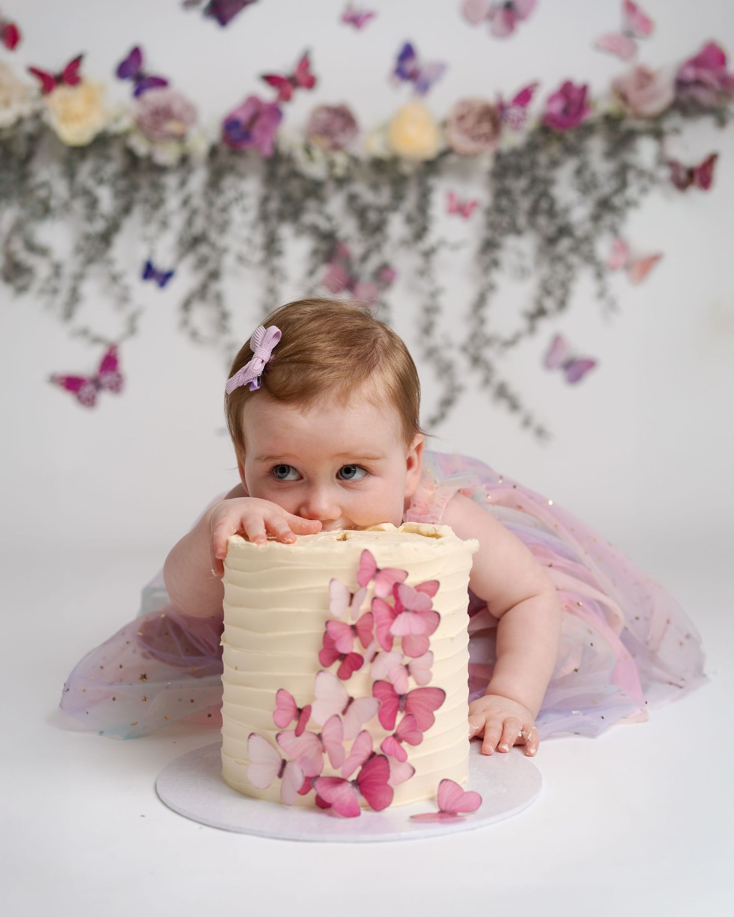 Baby girl in pink tutu biting butterfly-decorated first birthday cake, with floral and butterfly backdrop.