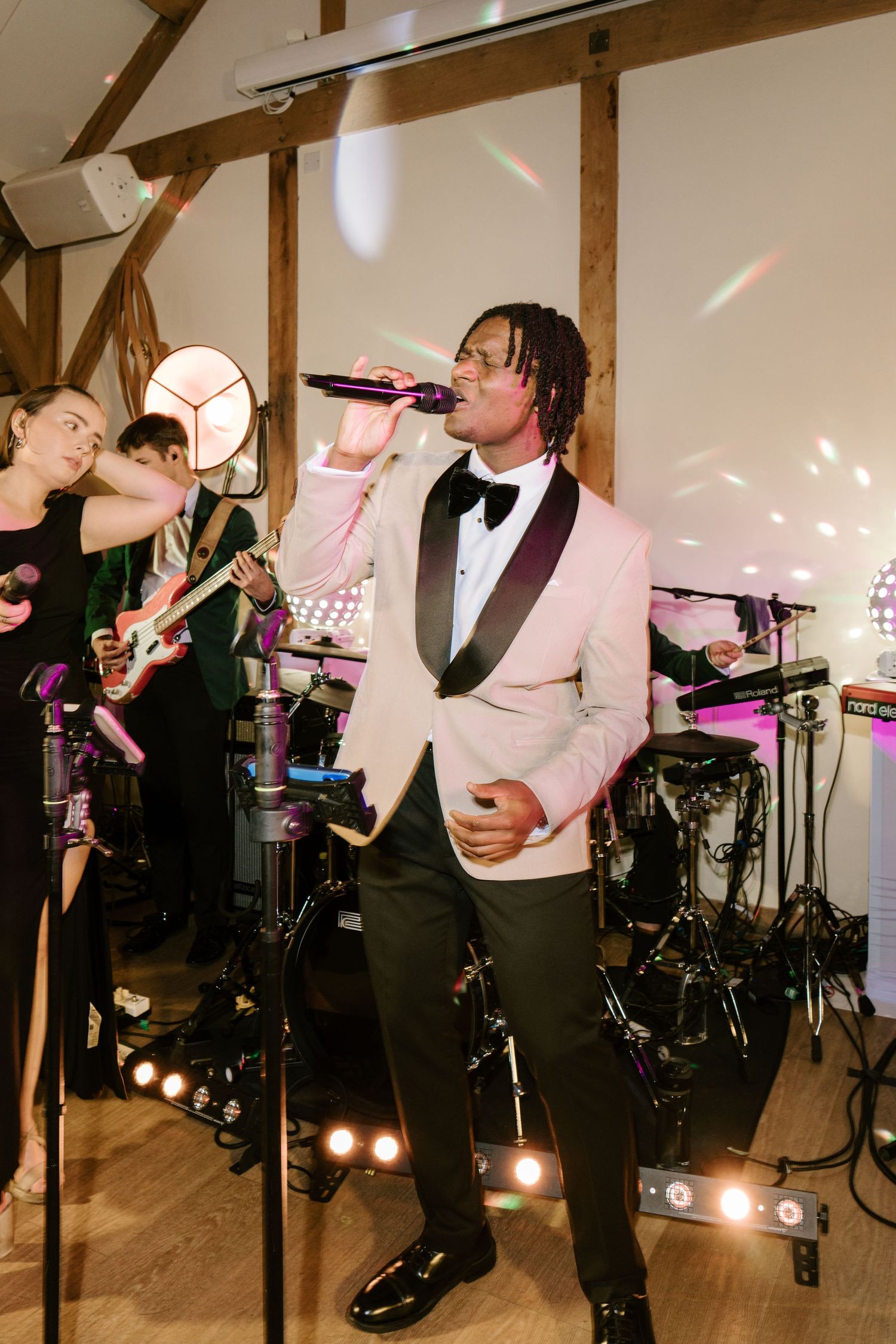 A male singer in a pink tuxedo jacket performs on stage with a live band in a warmly lit barn venue.
