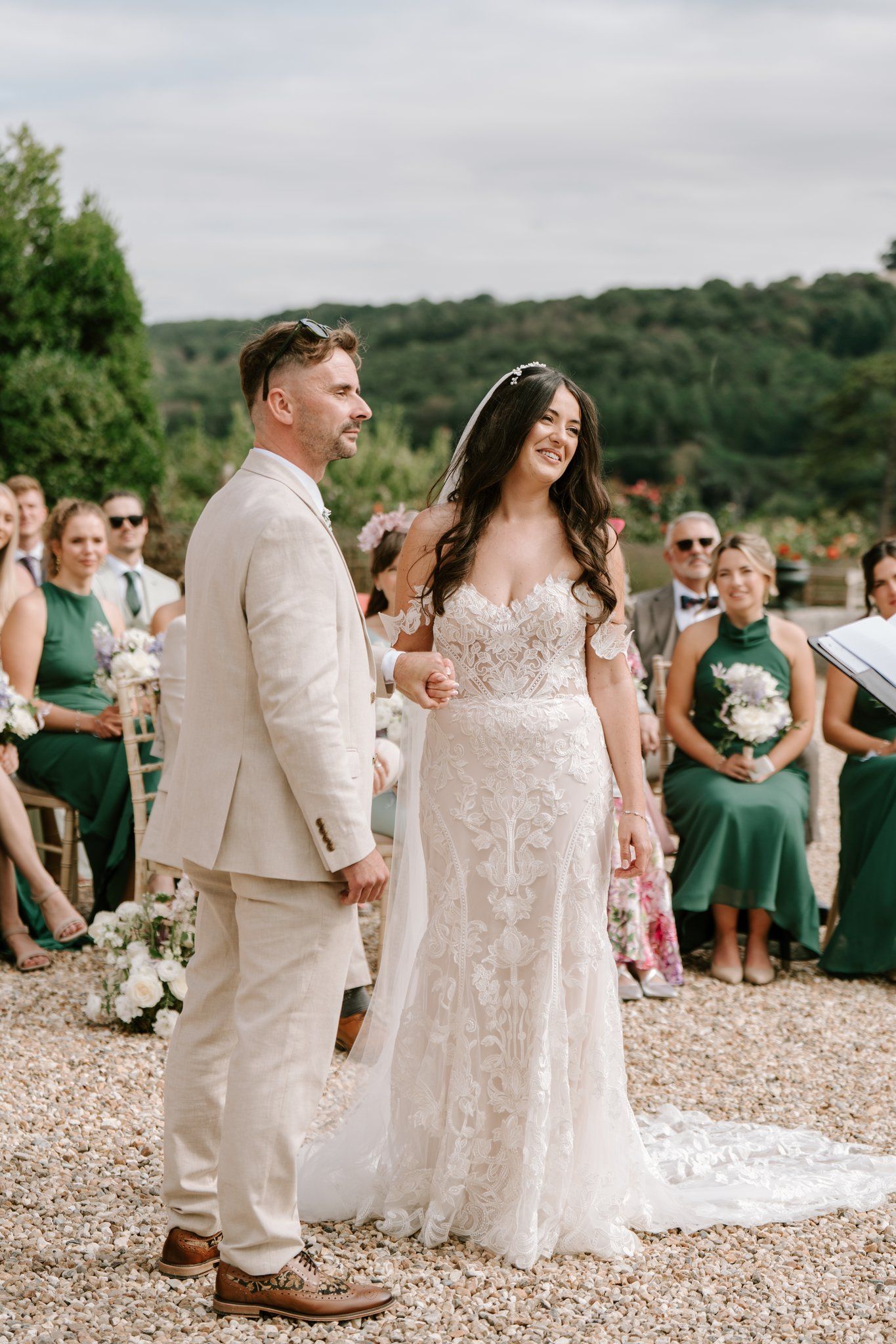 Bride in lace gown and groom in beige suit smile during outdoor wedding ceremony with guests in green attire.