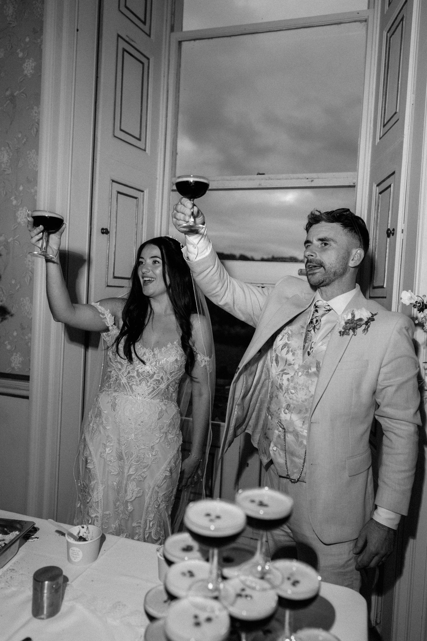 Black and white photo of bride and groom raising wine glasses in a toast at their wedding reception.