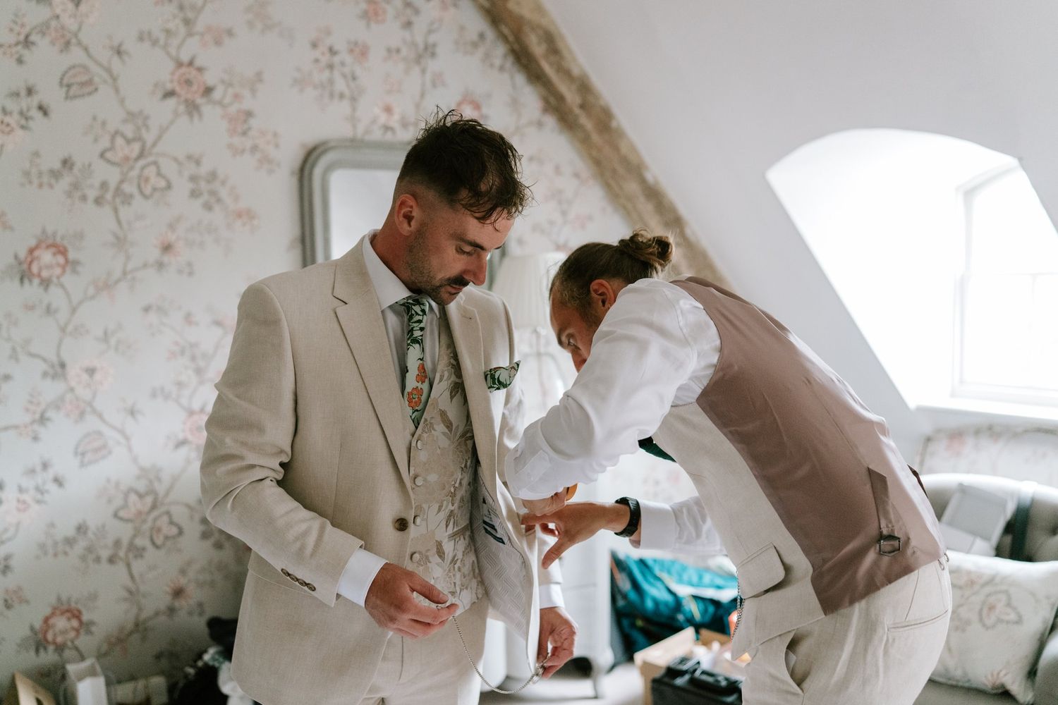 Groom in beige suit being helped by wedding assistant in bright floral wallpaper room on wedding day.