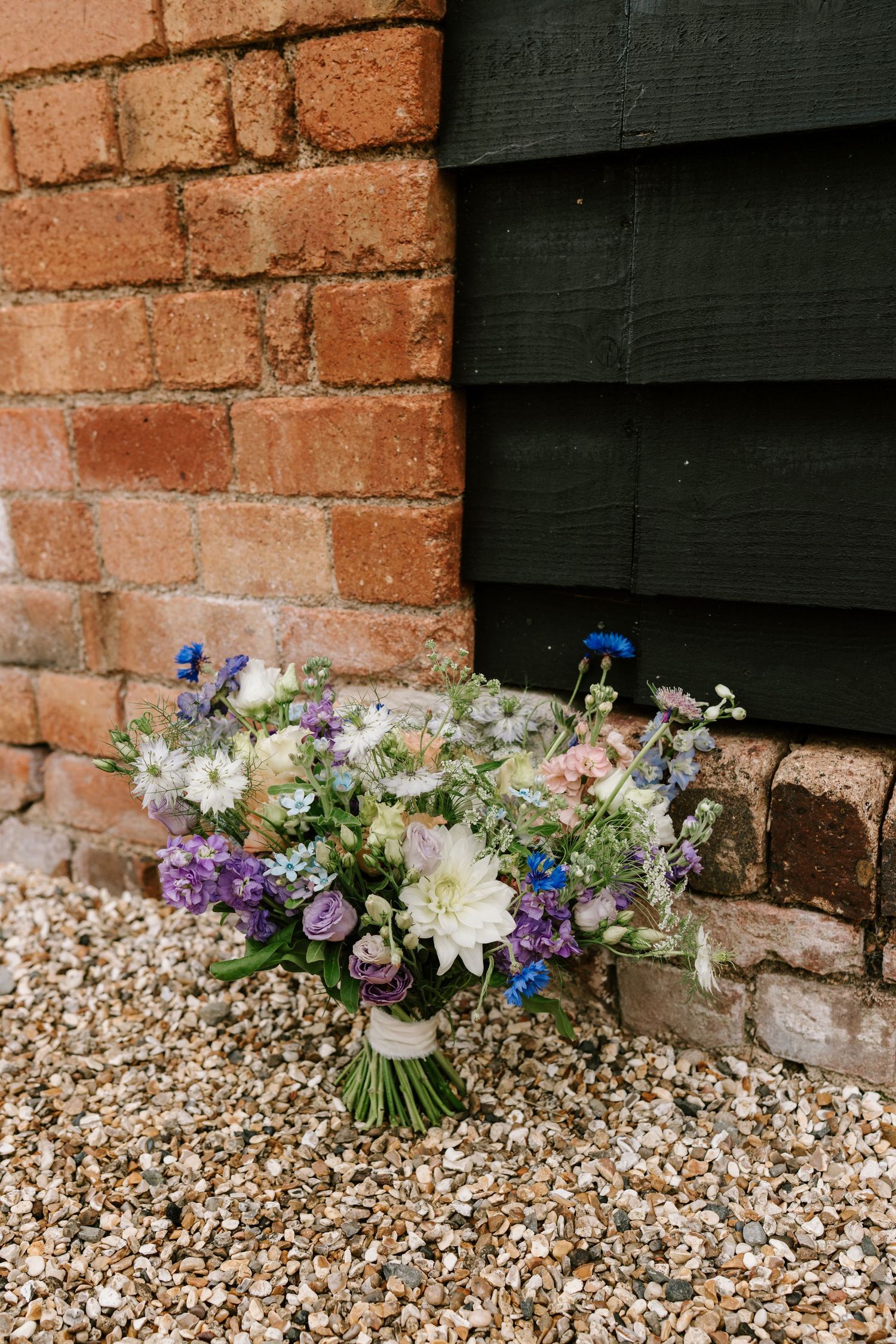 A wildflower wedding bouquet with purple, blue, and white blooms resting against a rustic brick wall on gravel.