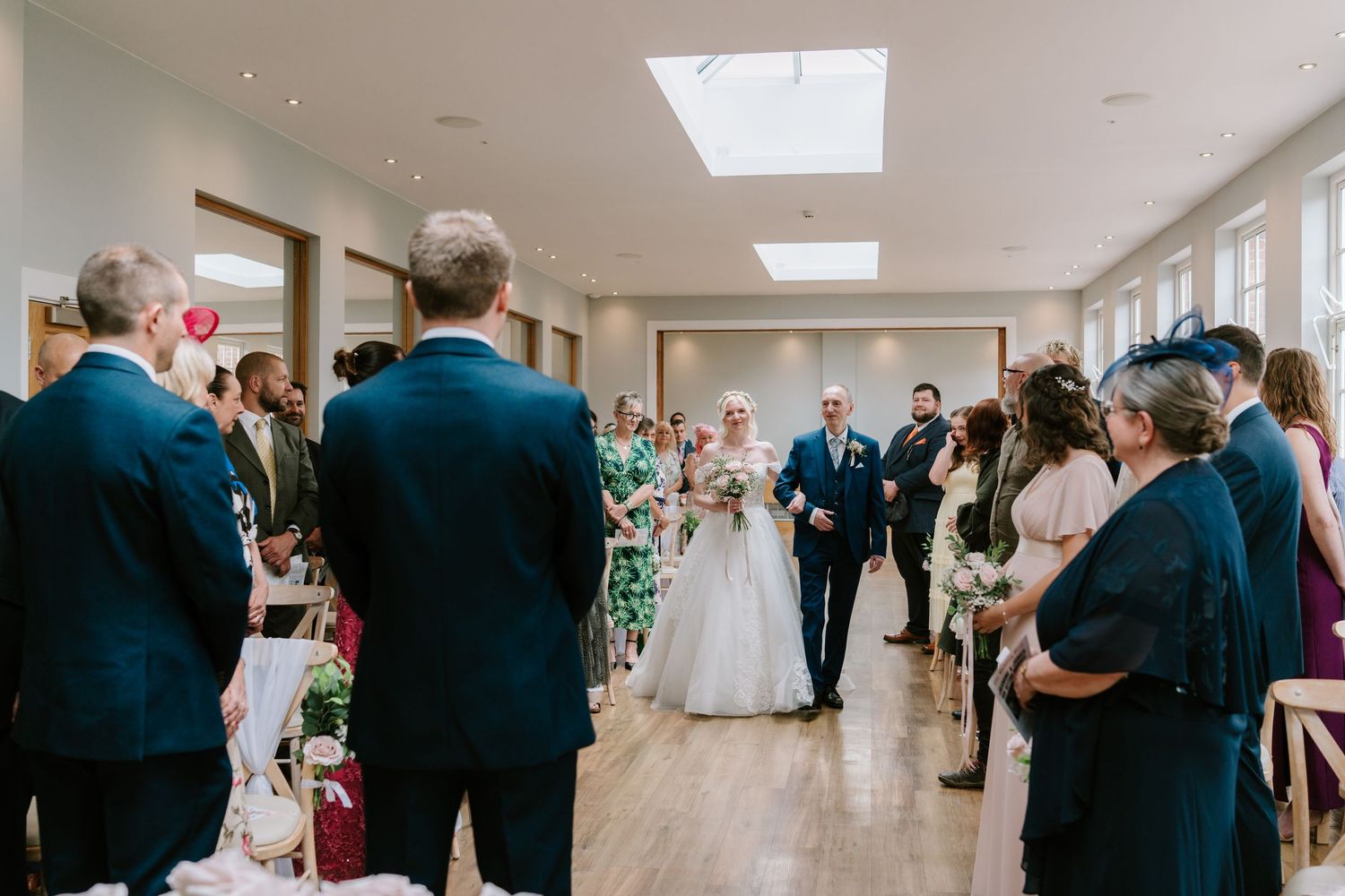 Bride walks down the aisle with her father during an elegant indoor wedding ceremony with guests standing.