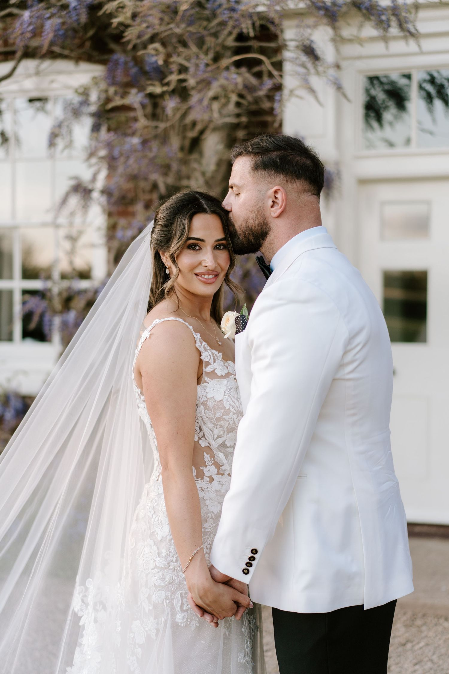 Bride in lace gown and veil holds hands with groom in white jacket as he kisses her cheek outdoors.