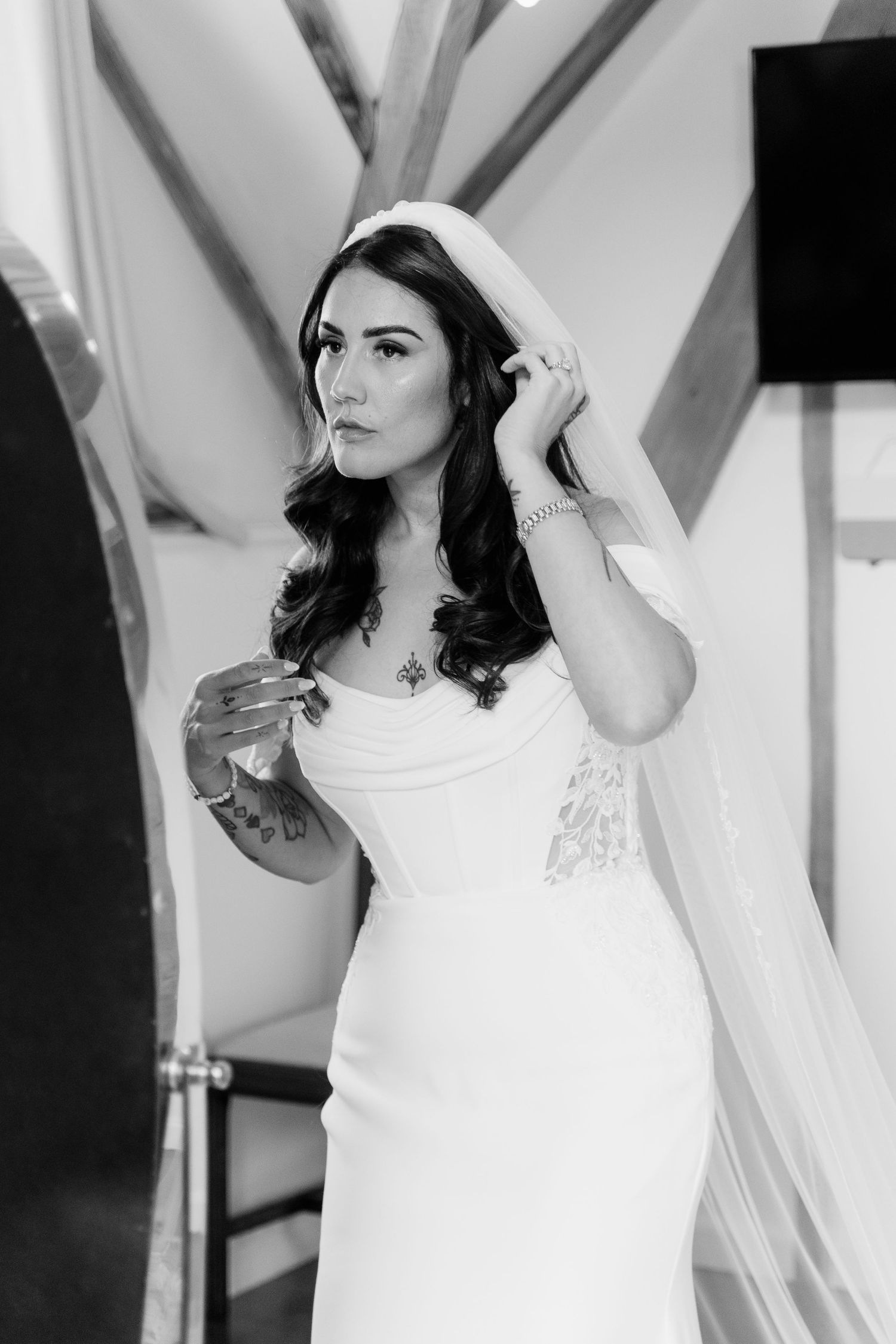 Black and white photo of a bride in a white gown and veil adjusting her hair, showcasing elegant bridal style.