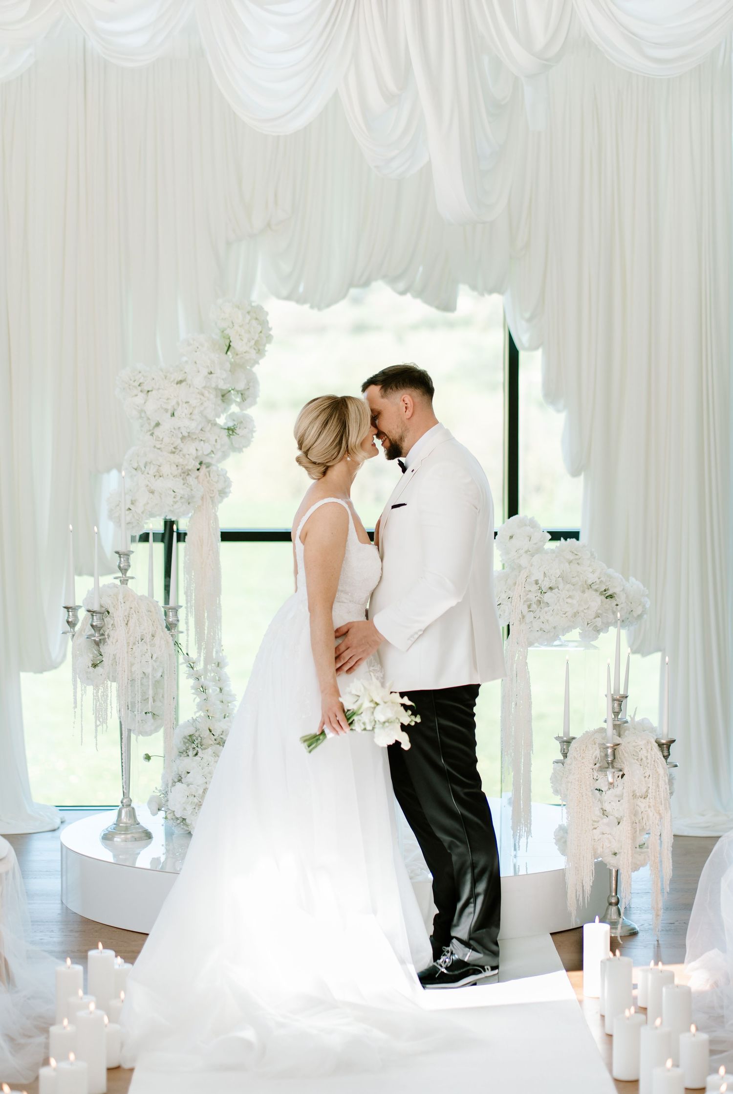 Bride and groom share a romantic kiss under a white floral arch surrounded by elegant candles and white draping.