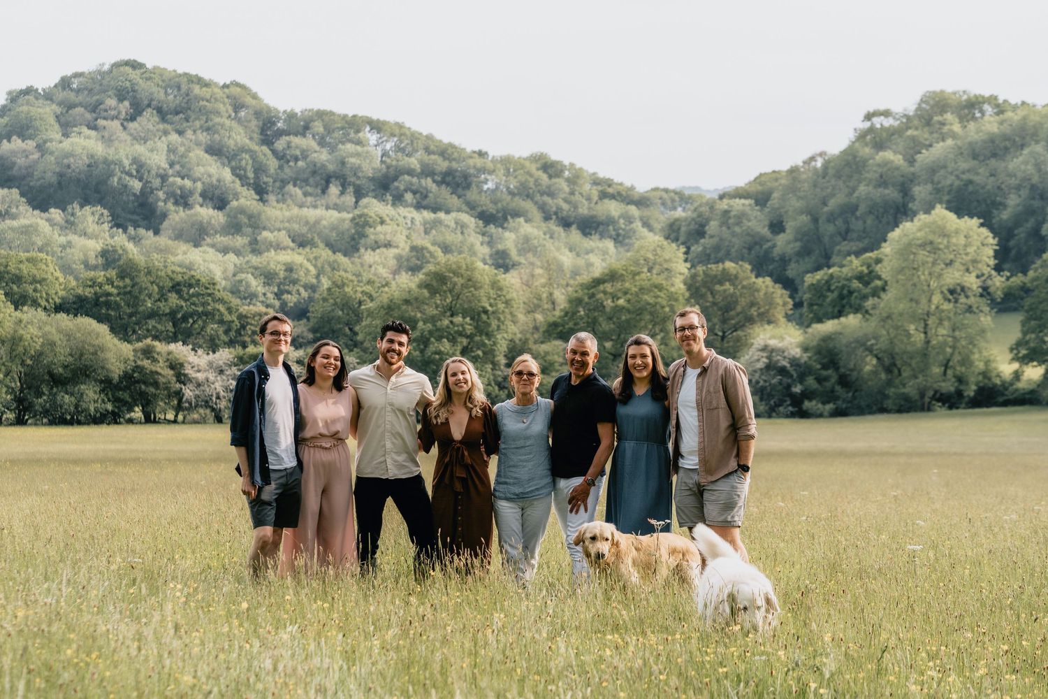 Group of seven people and a golden dog posing in a green meadow with tree-covered hills in the background.