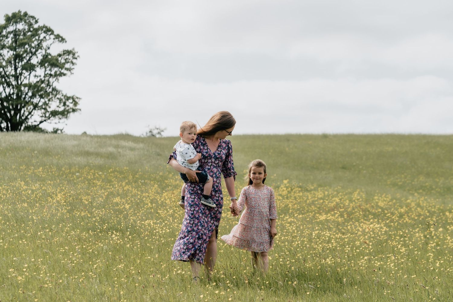 A mother walks through a sunny wildflower meadow holding a baby while a young girl in pink dress walks beside her.