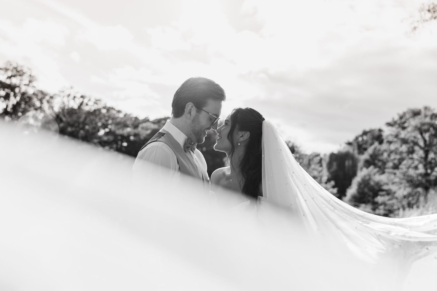 Black and white wedding photo of bride and groom embracing, veil flowing, surrounded by trees.
