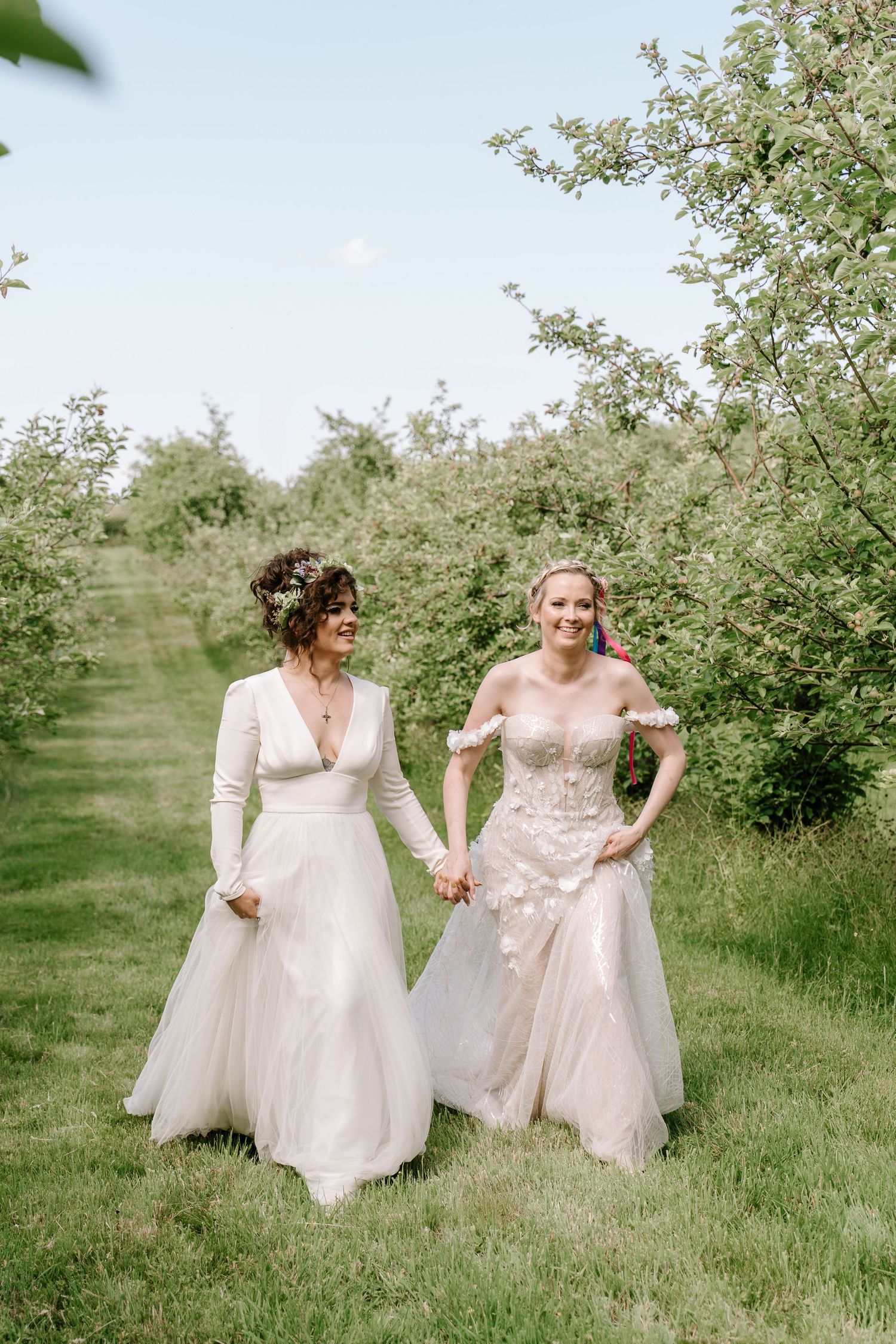 Two brides in white wedding gowns walk hand-in-hand through a lush green orchard on their wedding day.