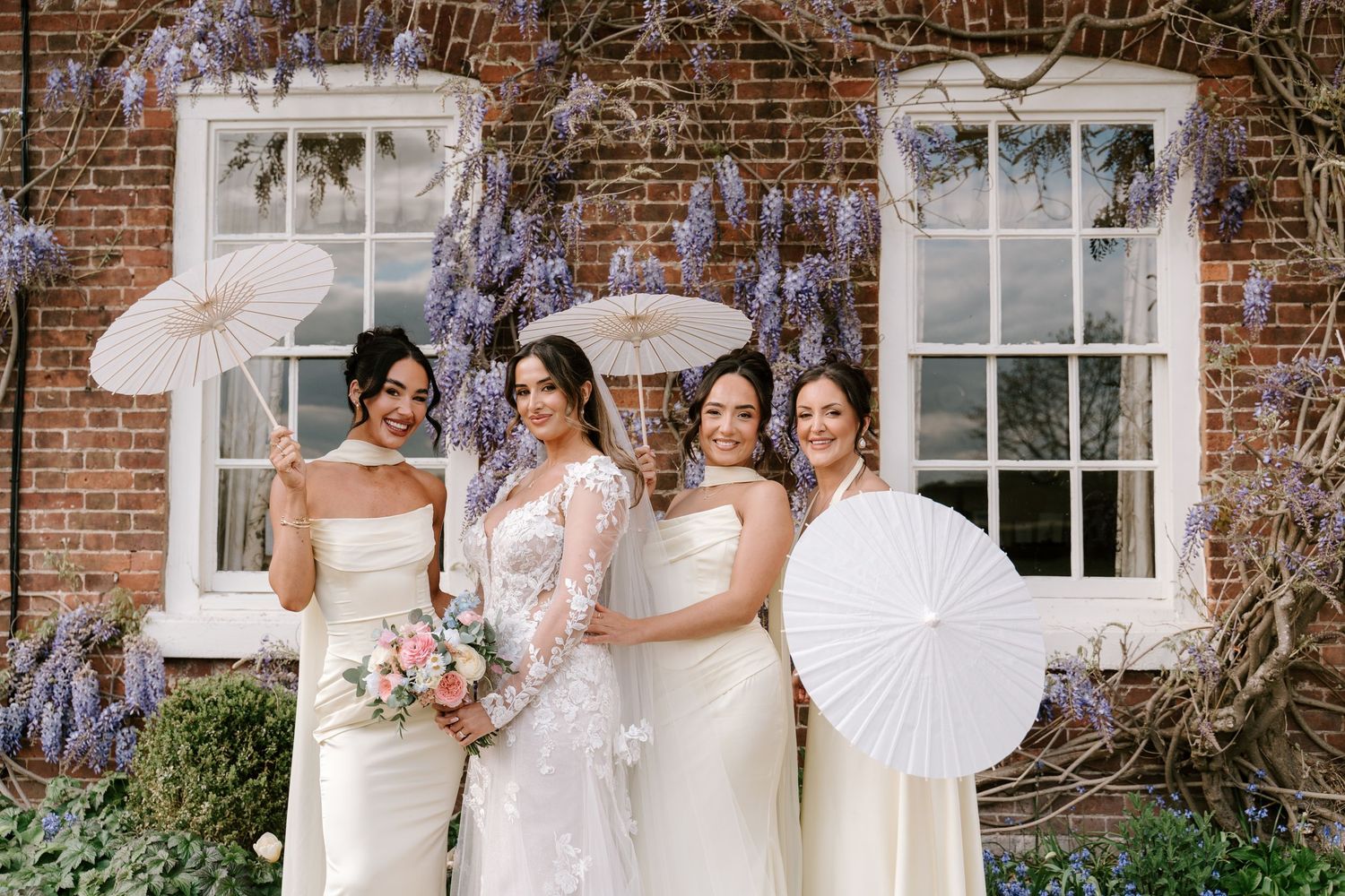 Bride and three bridesmaids in white gowns holding parasols pose before wisteria-covered brick wall.