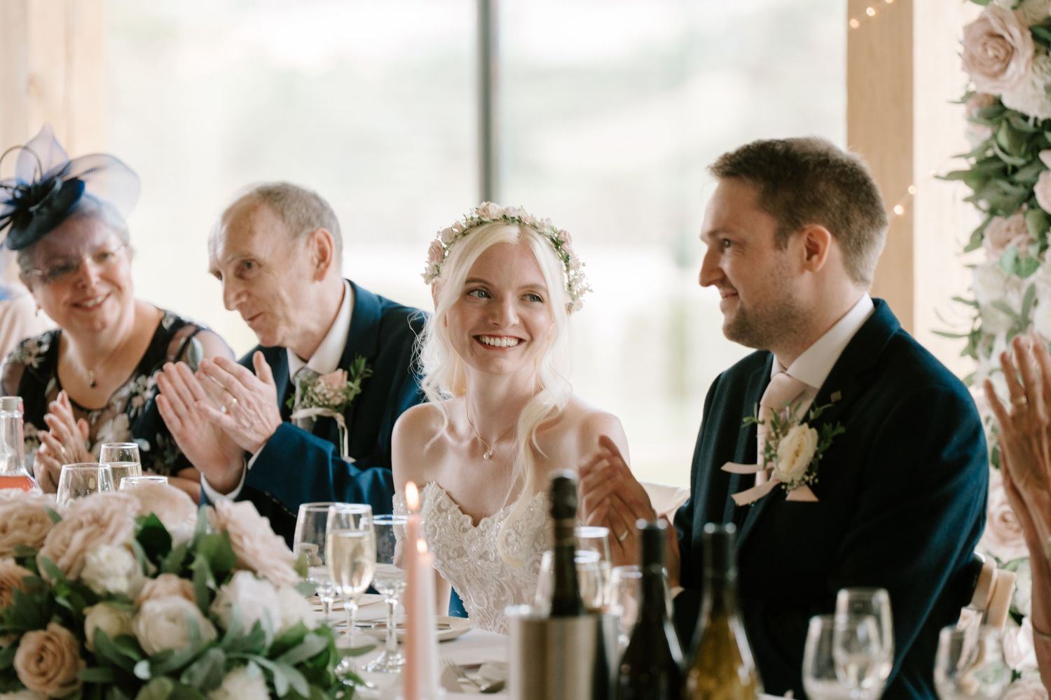 Bride and groom laughing at wedding reception table with family clapping in elegant floral venue.