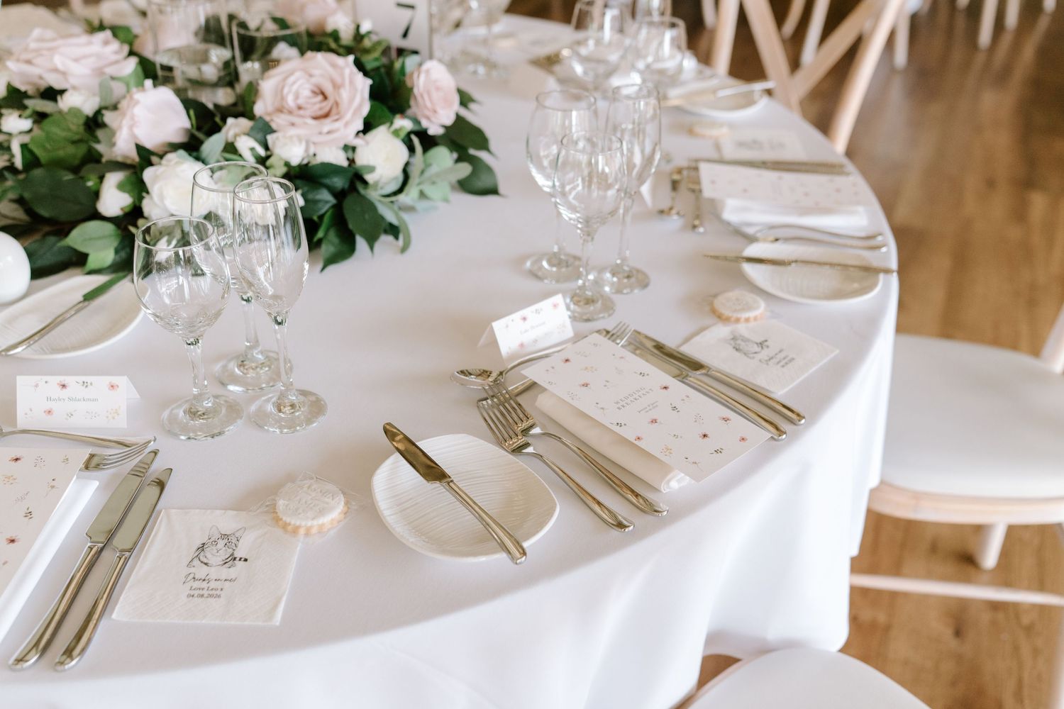 Elegant wedding table setting with white linens, blush roses, gold cutlery, crystal glasses, and delicate place cards.