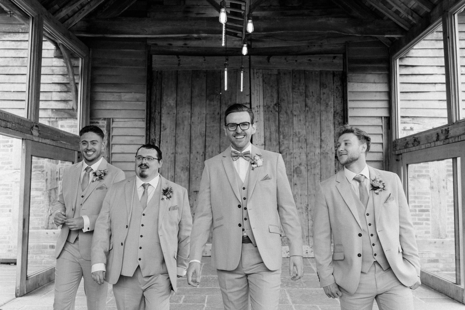 Black and white photo of groom and groomsmen in light suits posing in front of wooden barn doors at wedding.