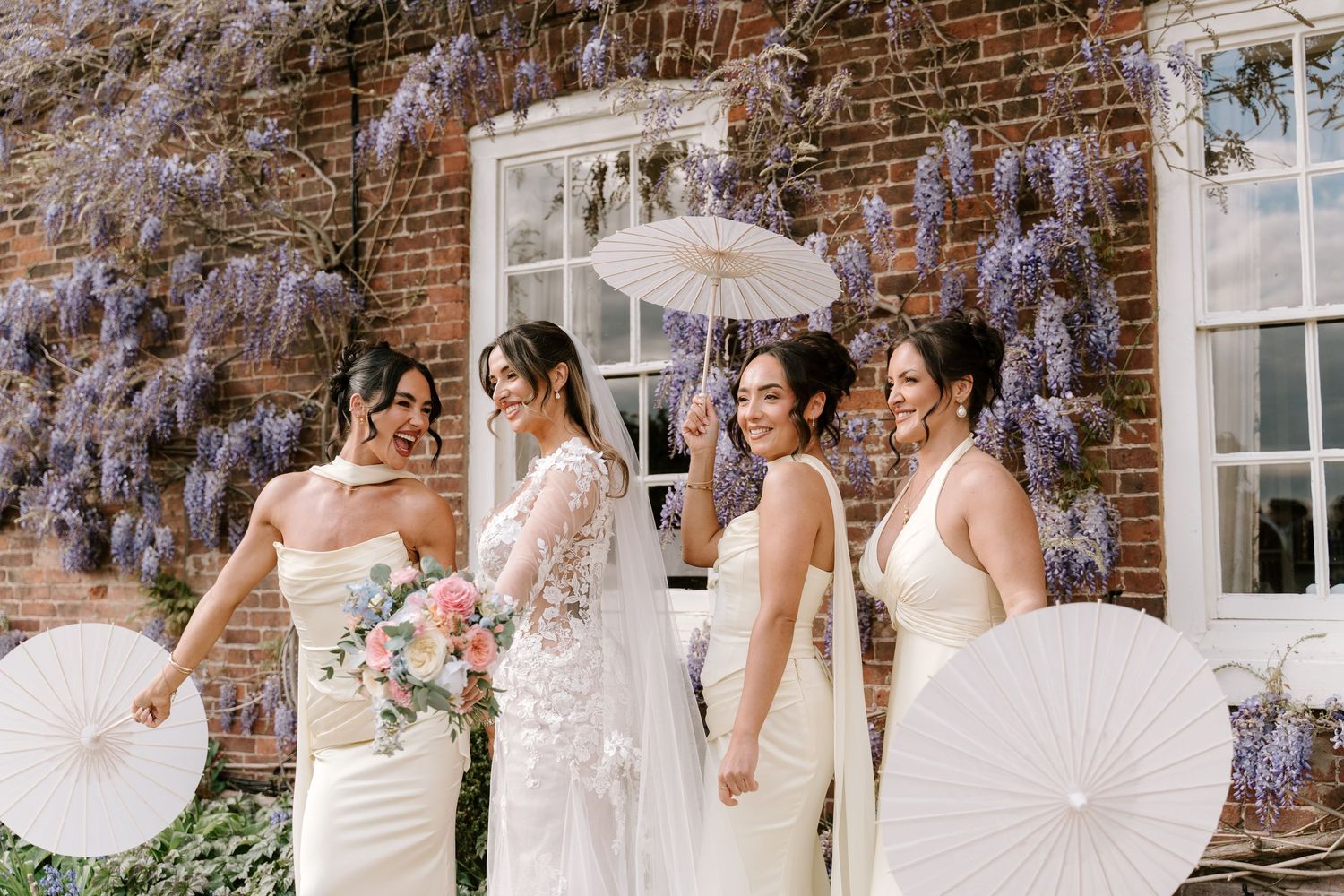 Bride and bridesmaids in white gowns holding paper parasols laugh together under blooming wisteria at elegant brick venue.