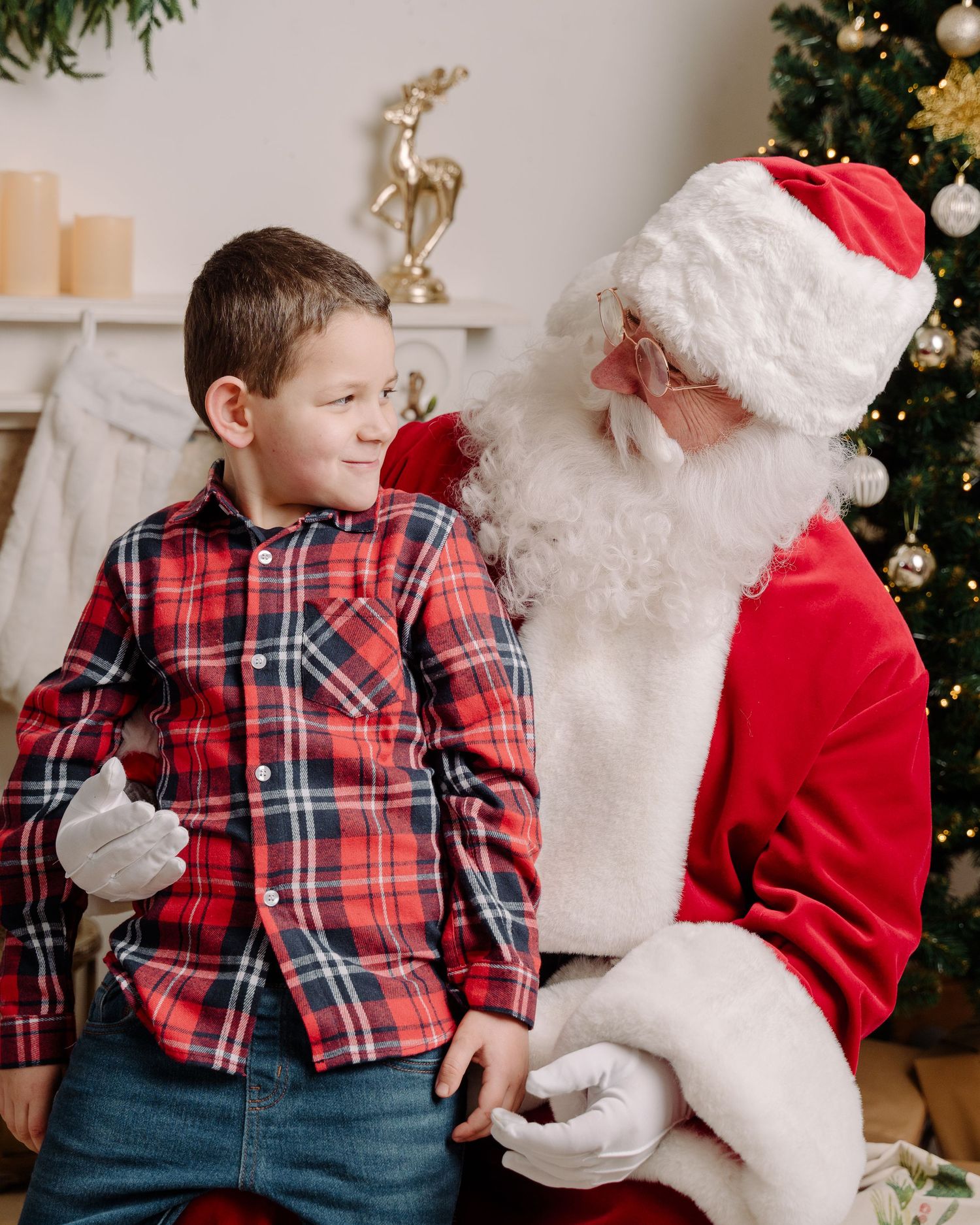 A young boy in a red plaid shirt sits with Santa Claus near a decorated Christmas tree and festive fireplace.