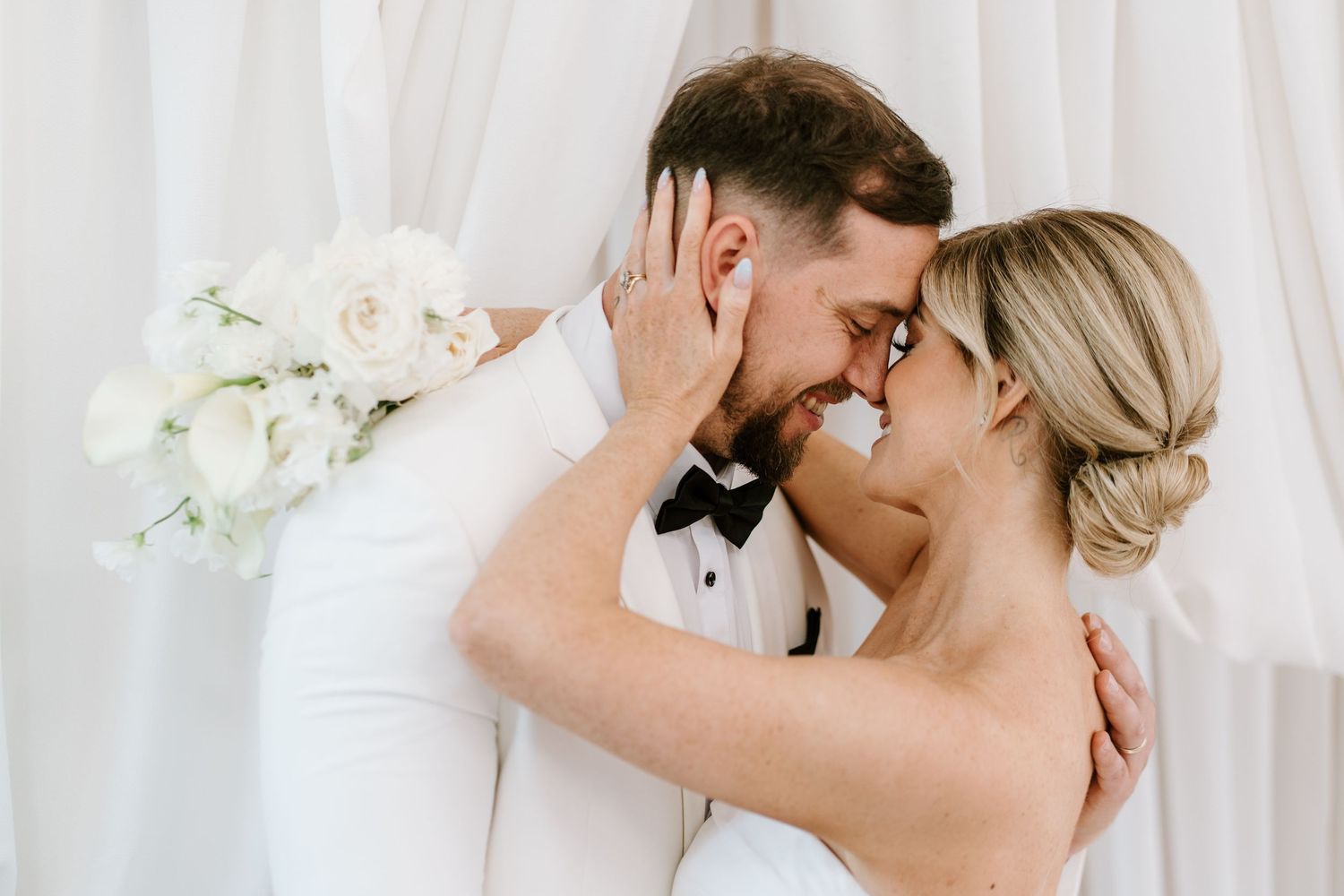 A bride and groom share a romantic forehead touch, she holds white roses, both dressed in elegant white wedding attire.