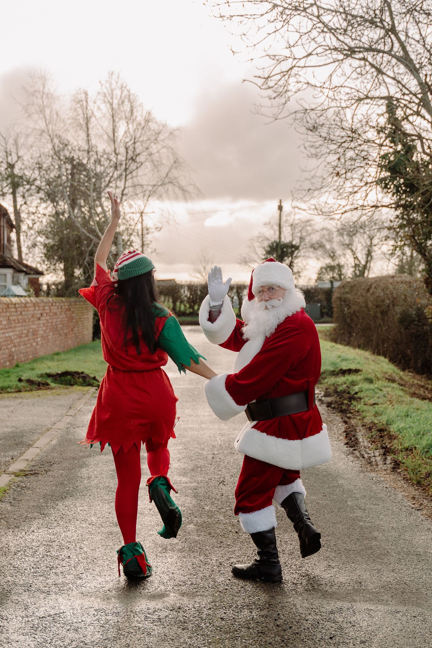Santa Claus and elf helper high-fiving on a rural road in festive Christmas costumes during winter.