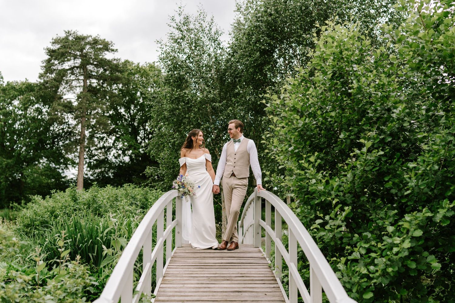 Bride and groom holding hands walking across white wooden bridge surrounded by lush greenery.