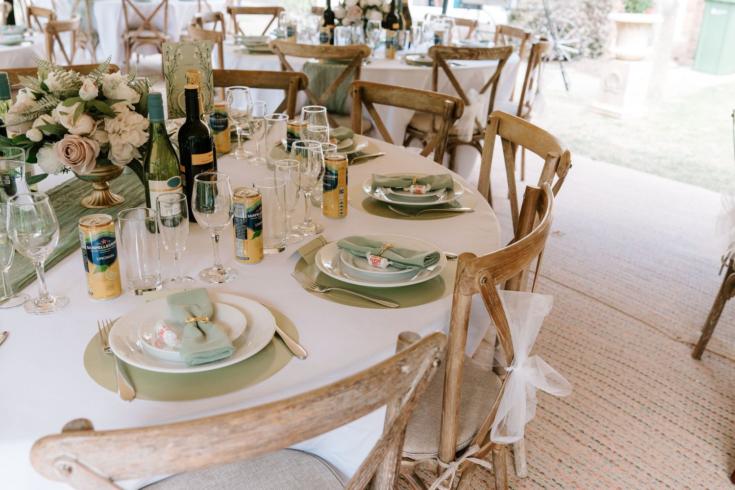 Elegant wedding reception table set with sage green plates, wine bottles, and blush floral centerpiece on white linen.