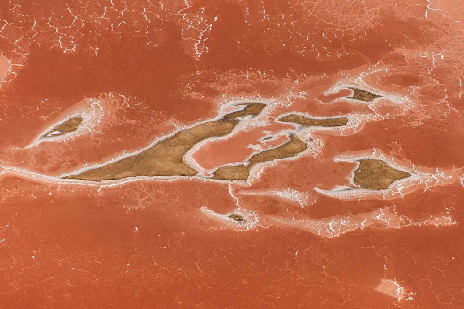 Aerial view of red salt pan with sandy island formations and white mineral deposits.