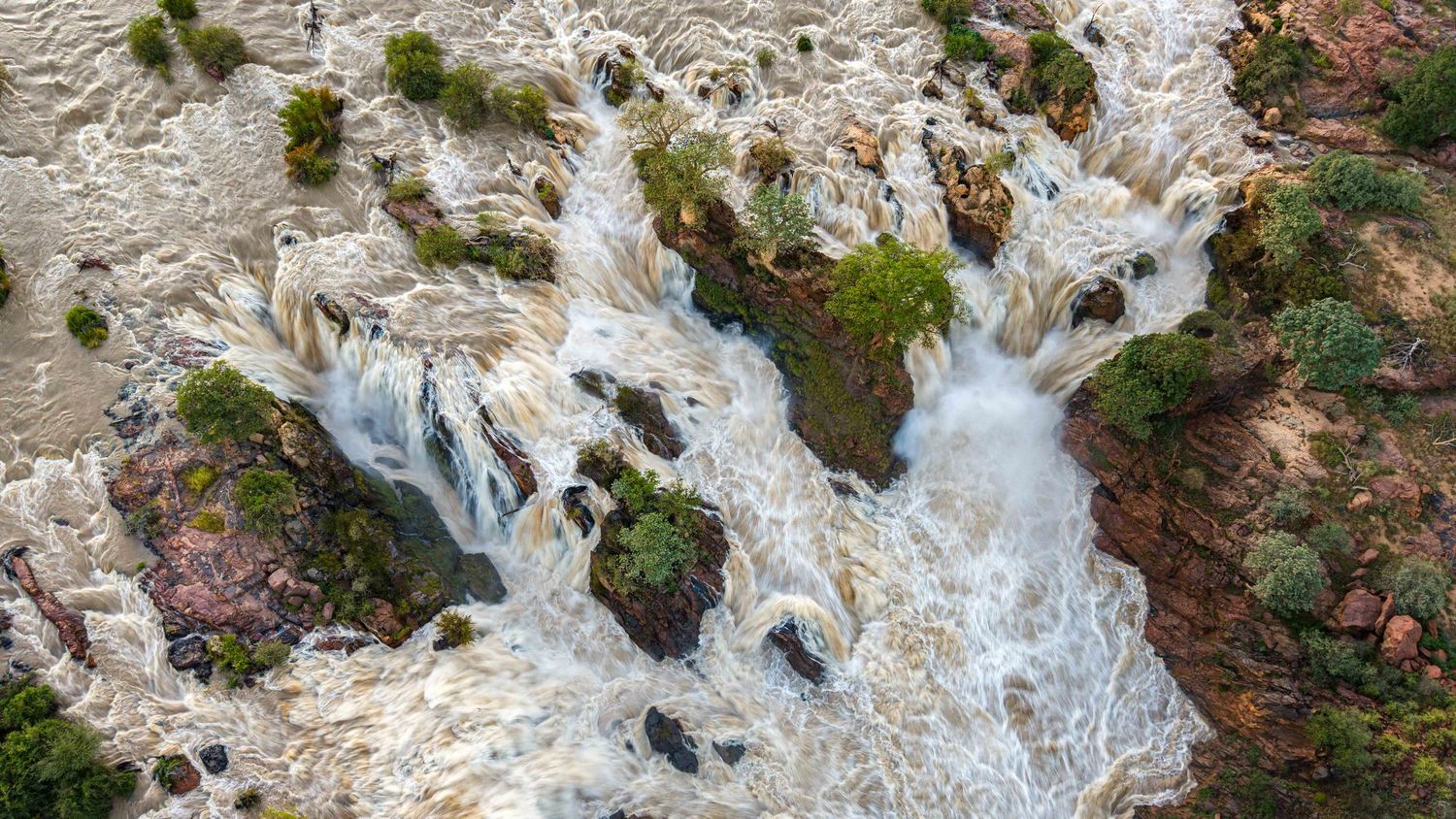 Bird's eye view of Epupa Falls showing powerful white water rushing over red rocks and green vegetation.