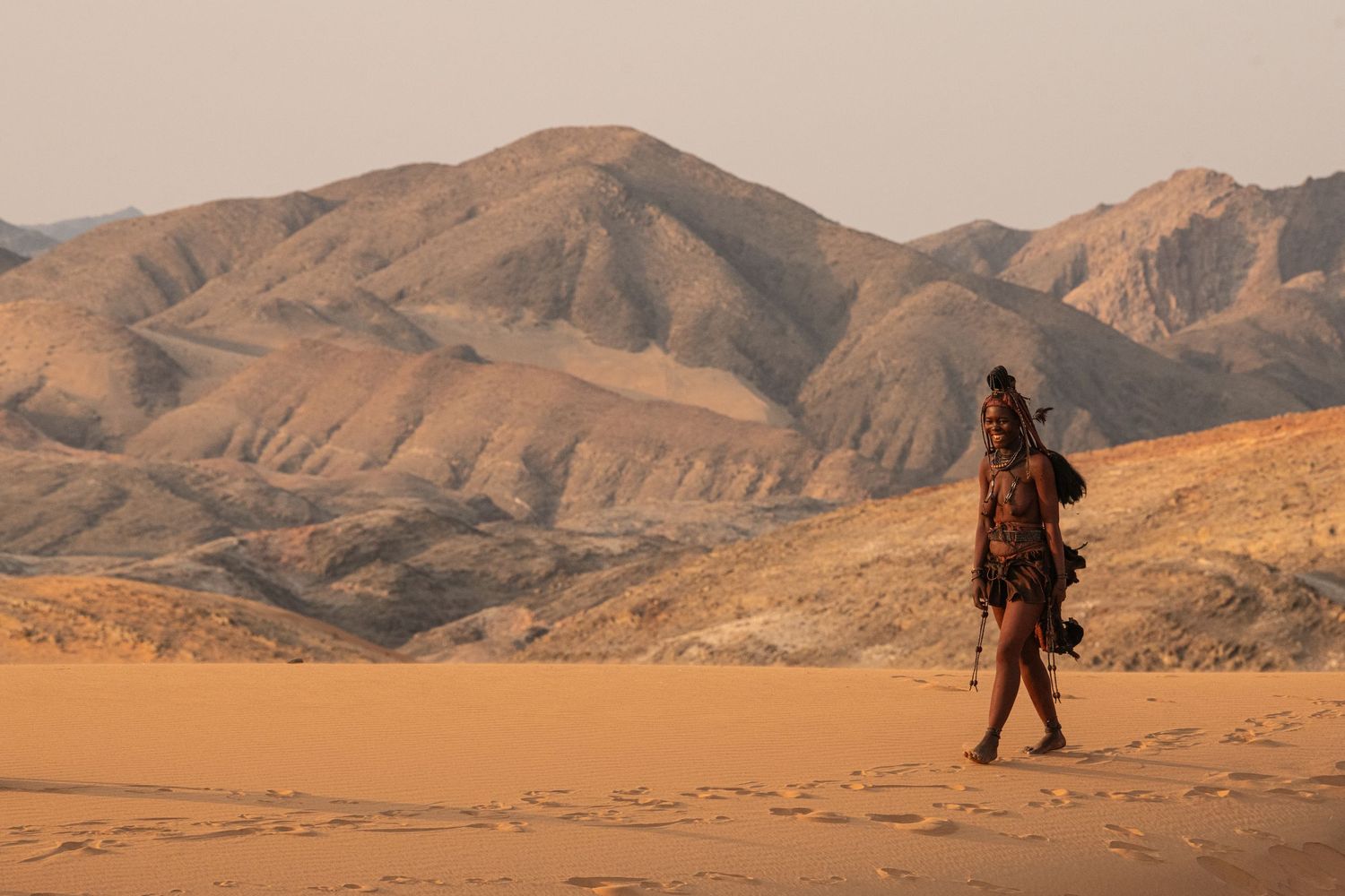 Himba tribe woman walking alone across vast orange sand dunes with dramatic mountain scenery.