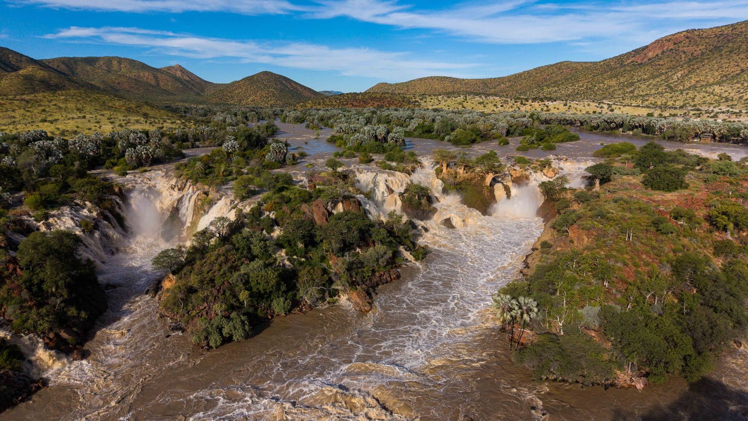 Aerial view of Epupa Falls with rushing water flowing through rocky terrain surrounded by green hills.