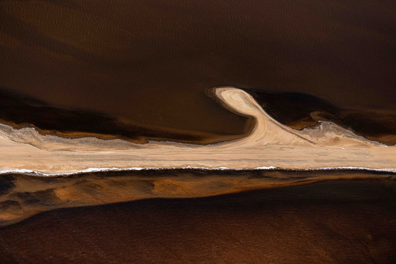 Aerial abstract view of dark brown sand dunes with a winding white dry riverbed in desert.