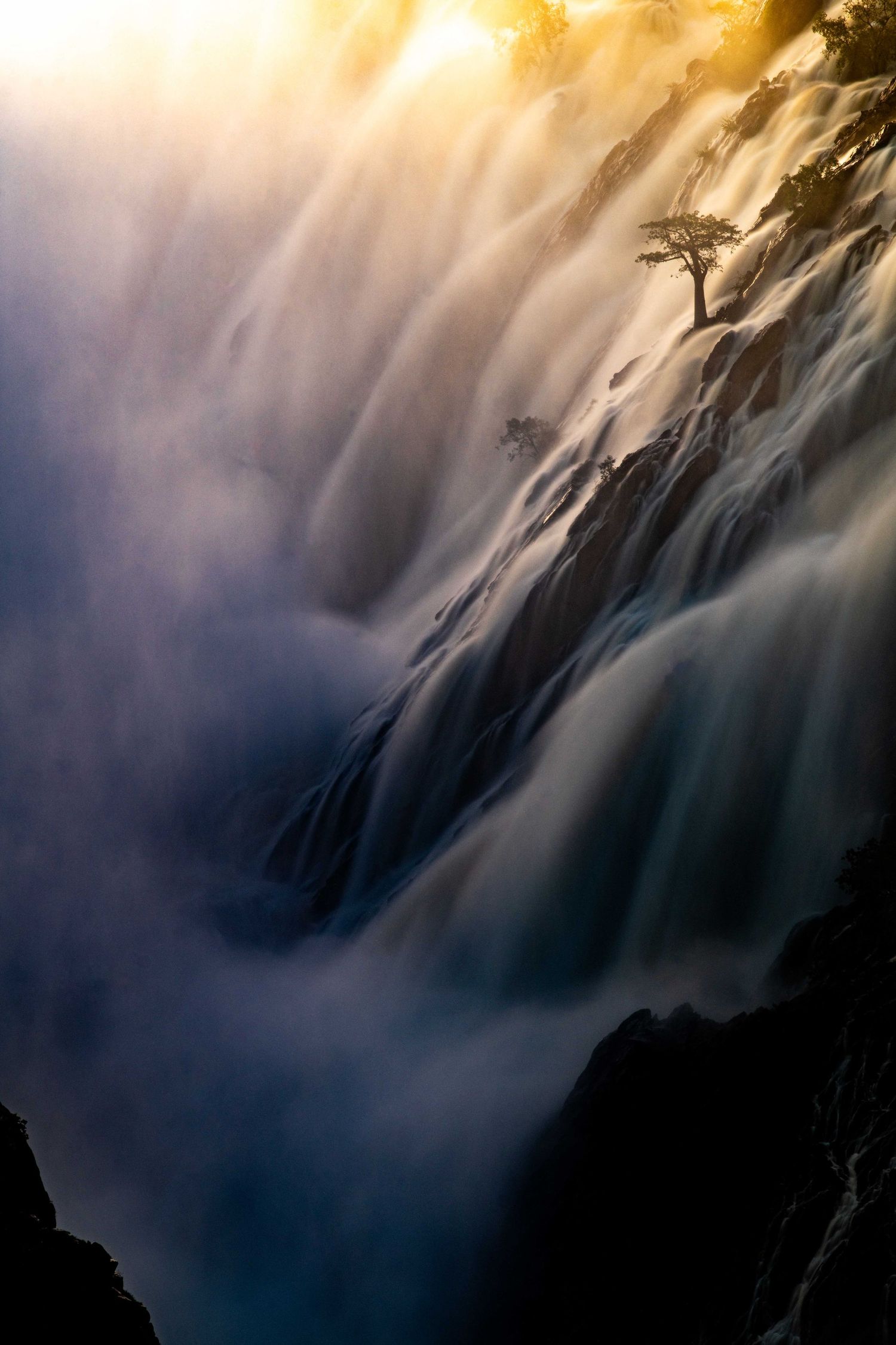 Powerful waterfall cascading over rocky cliffs with a lone tree silhouetted against golden misty light.