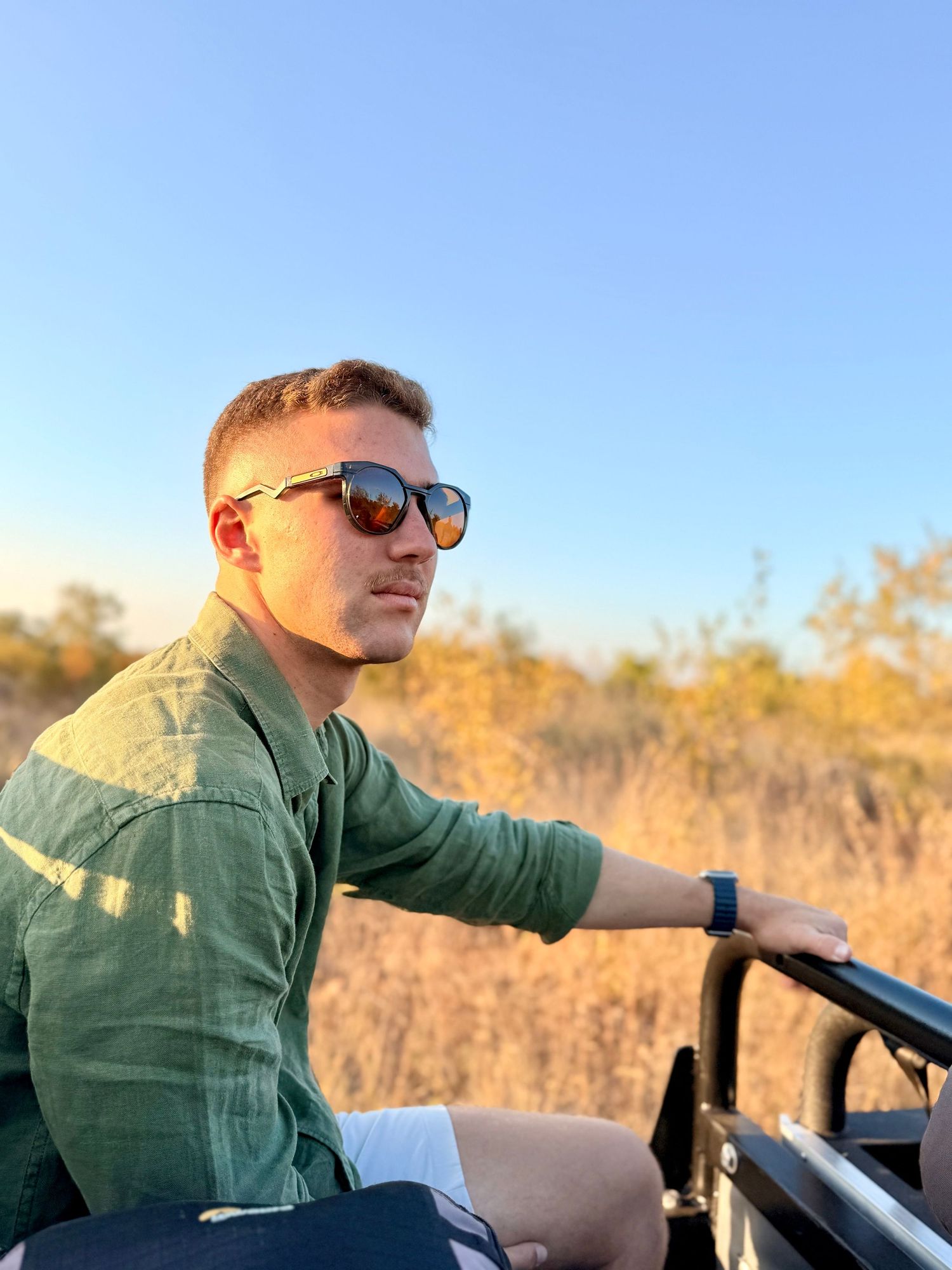 Close-up of man wearing sunglasses on safari vehicle at sunset with golden African bushveld in background.