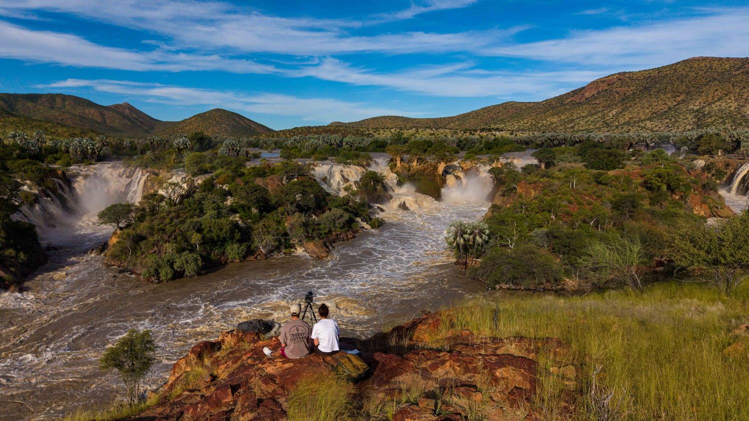 Two people sitting on red rocks overlooking the scenic Epupa Falls waterfall in Namibia under blue sky.