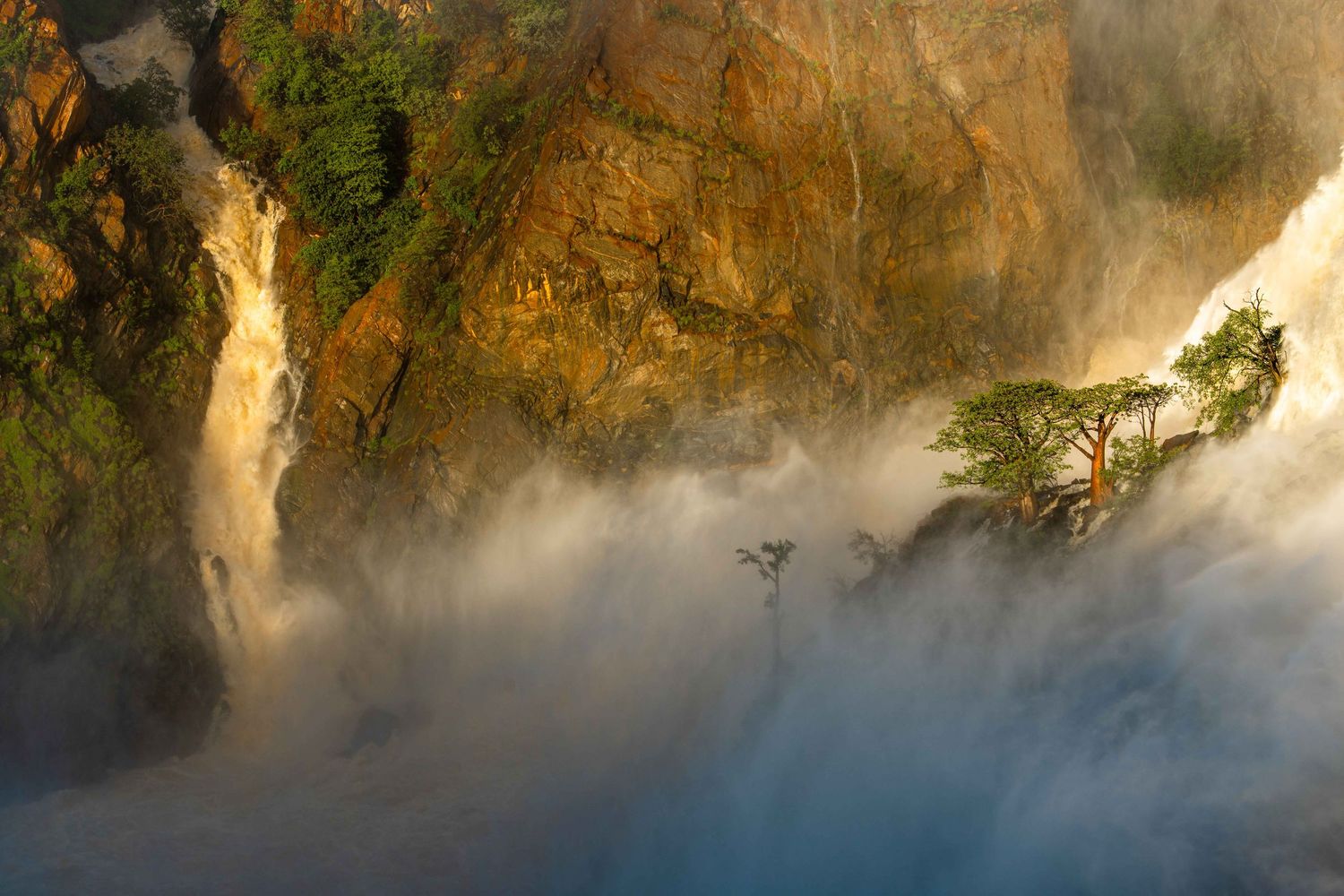 Misty waterfall cascading over golden rocky cliffs with lush green trees emerging from the fog.