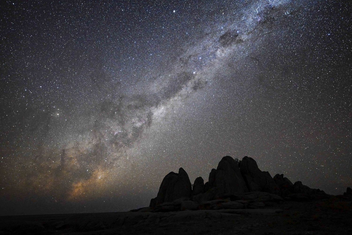 Milky Way galaxy arching over silhouetted rock formations in a dark desert night sky filled with stars.