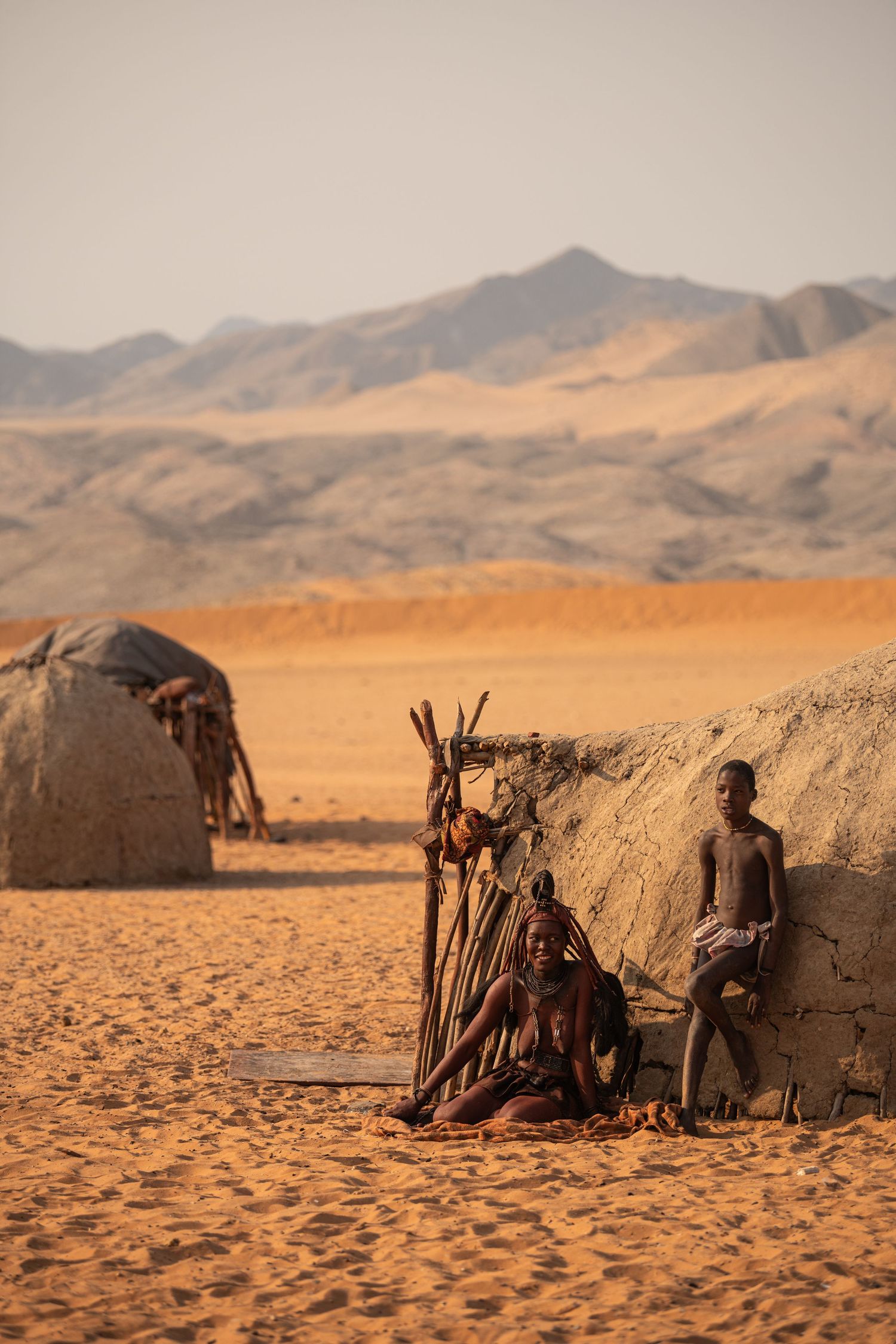 Himba tribe woman and child resting beside a traditional mud hut in the Namibian desert landscape.