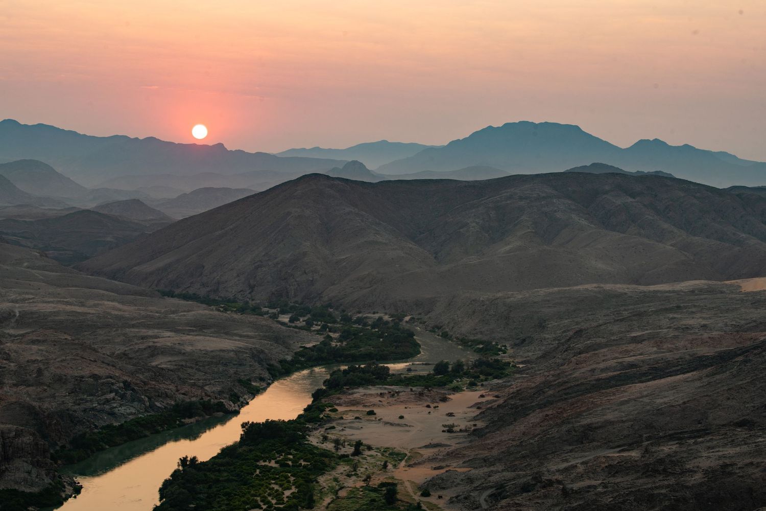 Sunset over winding river valley surrounded by layered mountain silhouettes in Namibia.