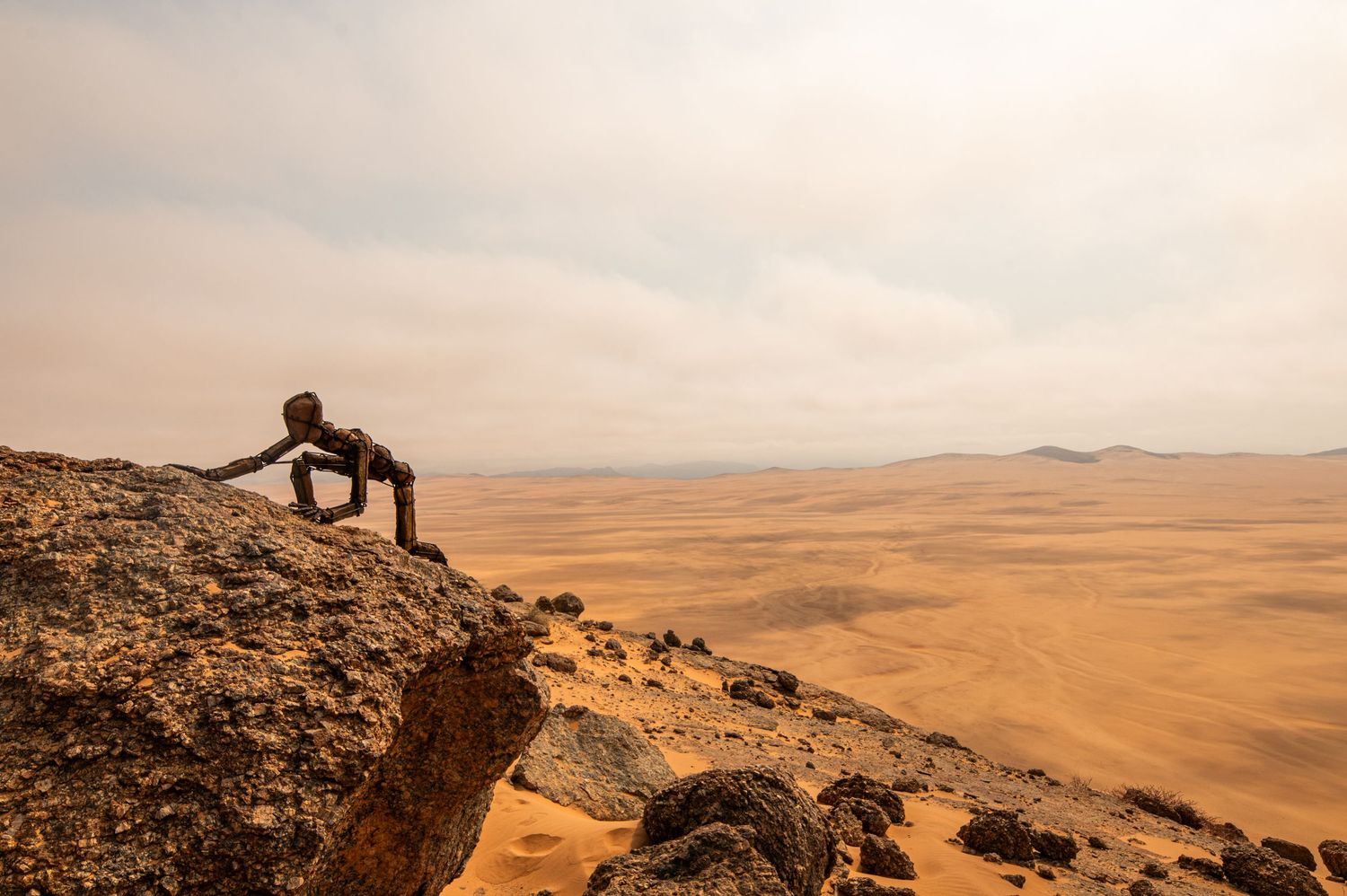 Metal sculpture of a figure peering over rocky cliff edge overlooking an expansive orange desert.
