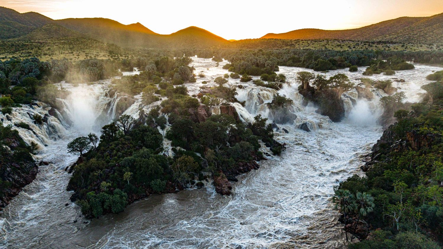 Aerial view of Epupa Falls on the Kunene River at sunset, with rushing white water and lush green trees.