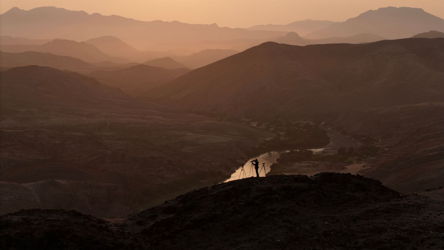 A silhouetted person stands on a rocky peak at sunset over a vast mountain landscape.