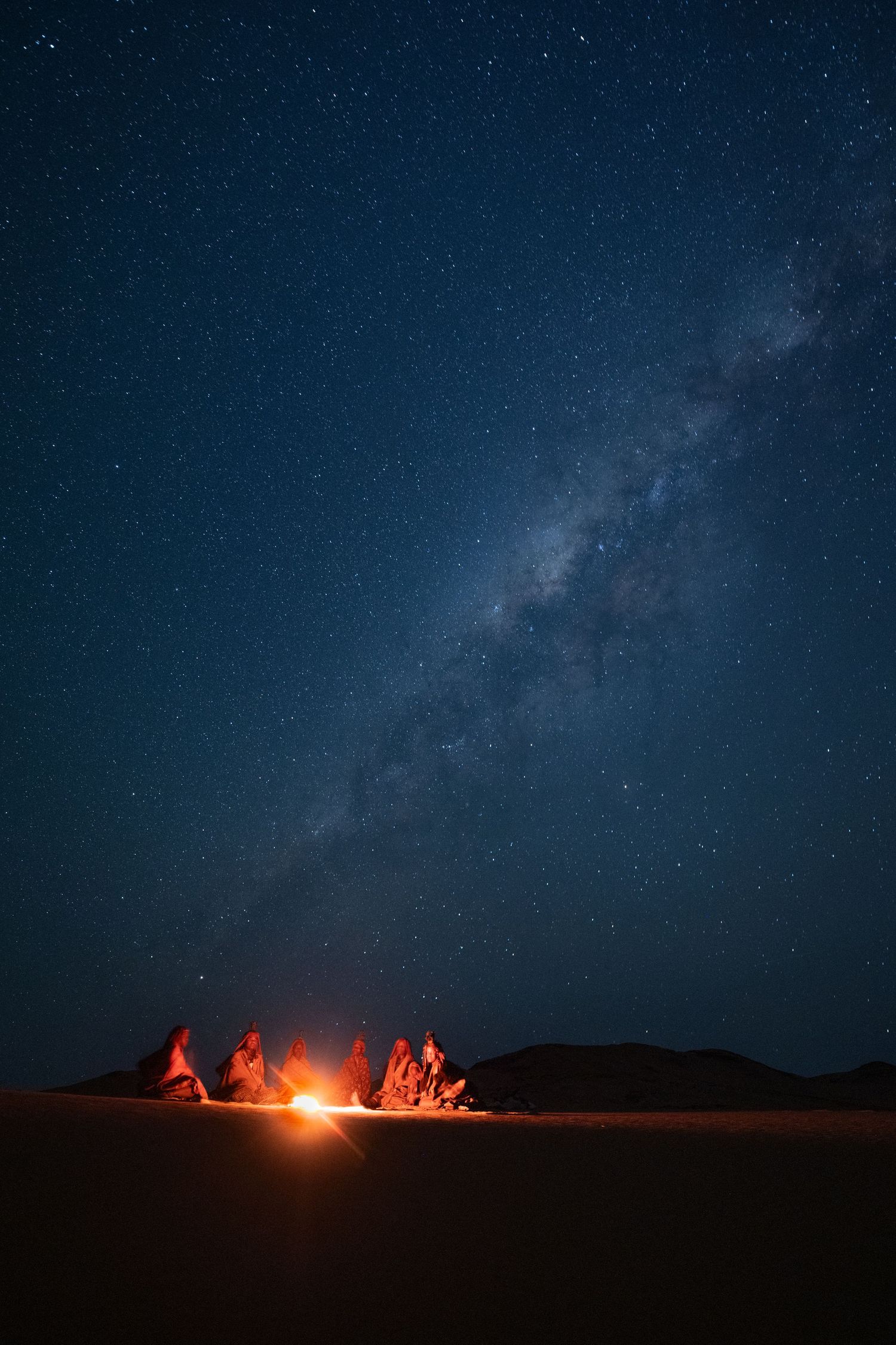 Campfire glowing under a stunning Milky Way night sky in a vast desert landscape.