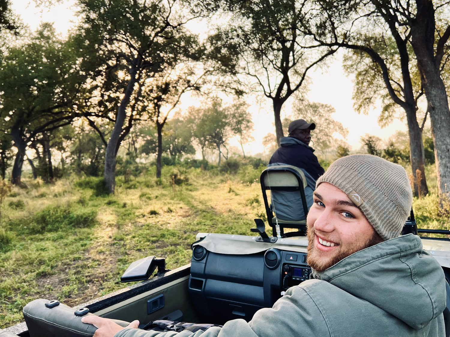A smiling man in a beanie sits in a safari vehicle at golden hour in the African bush.
