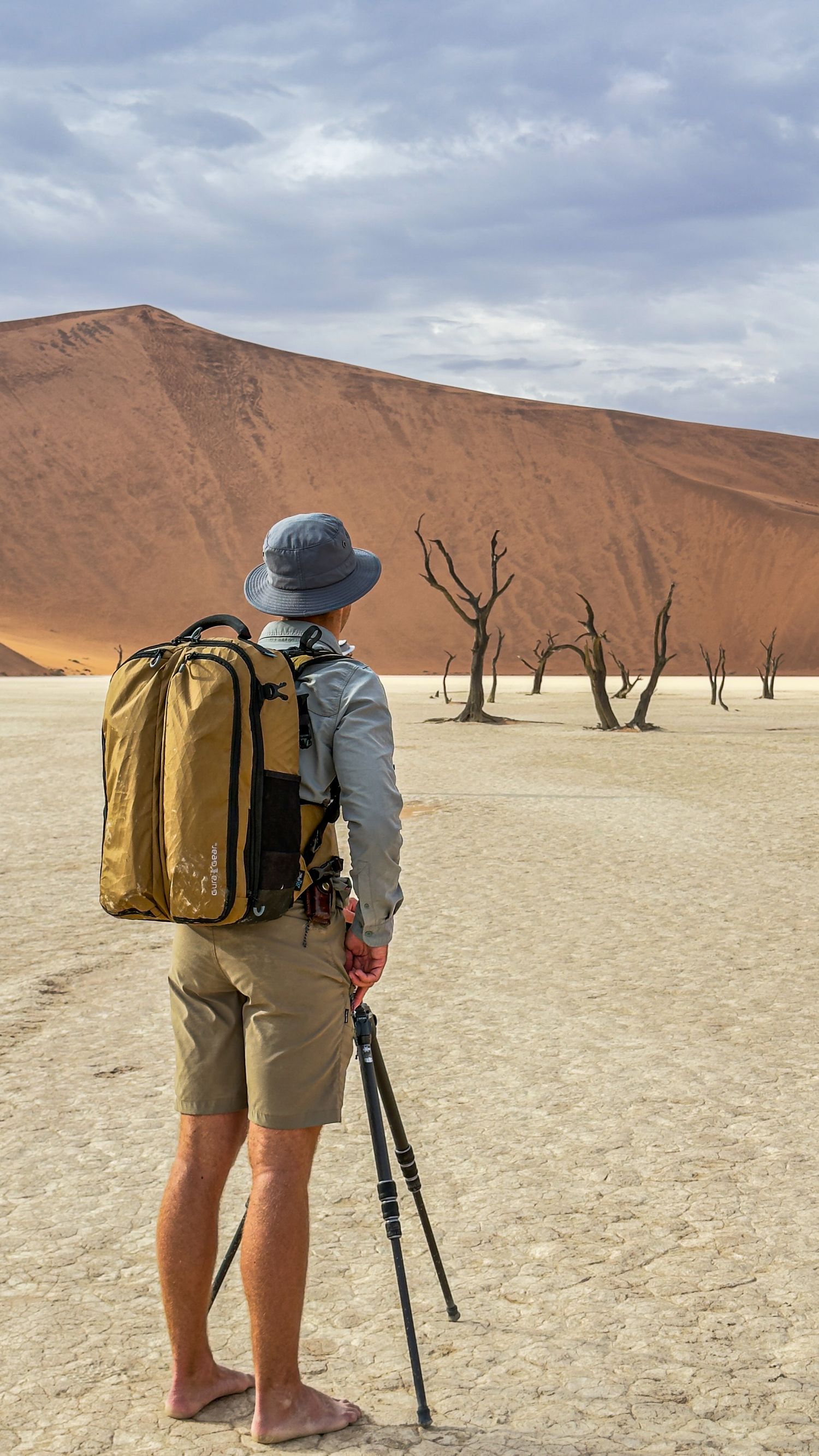 A photographer with backpack and tripod stands before dead trees and red dunes in Deadvlei, Namibia.