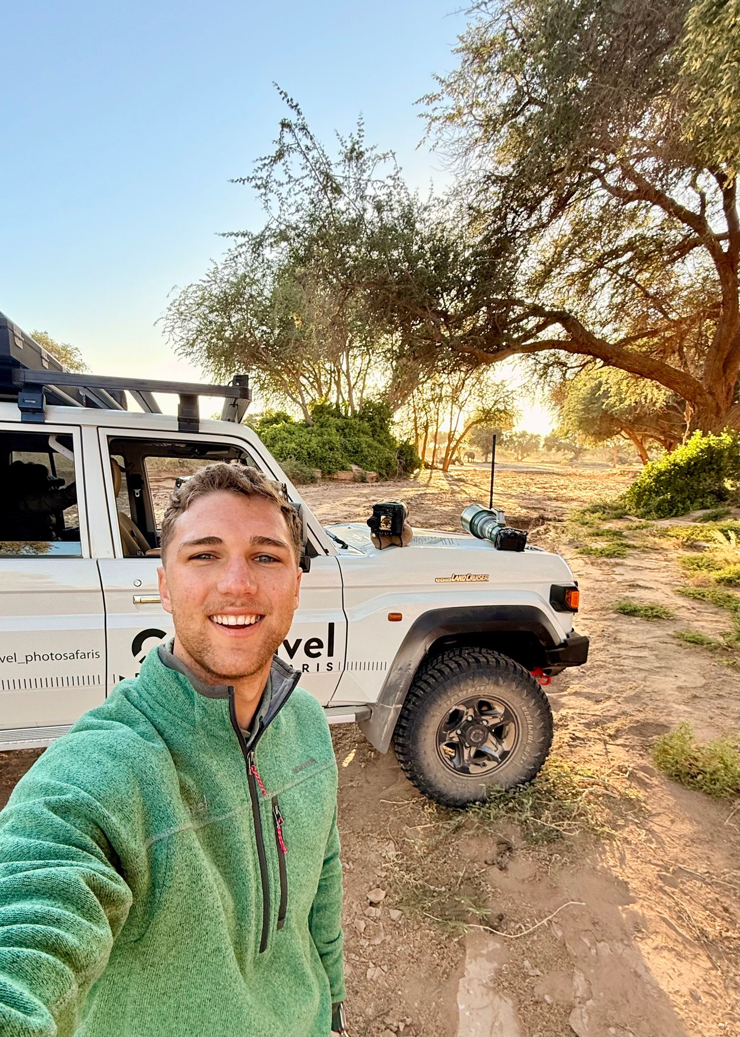 Man in green fleece takes selfie with safari jeep under acacia tree at golden hour in African wilderness.